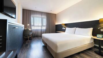 A hotel room with a large bed, a desk with a chair, a television mounted on the wall, and a window with curtains.