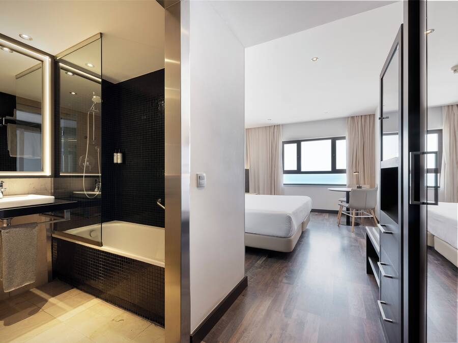 A modern hotel room with a bathroom featuring a bathtub, a large mirror, and a shower. The room has a bed, a desk, and a TV.