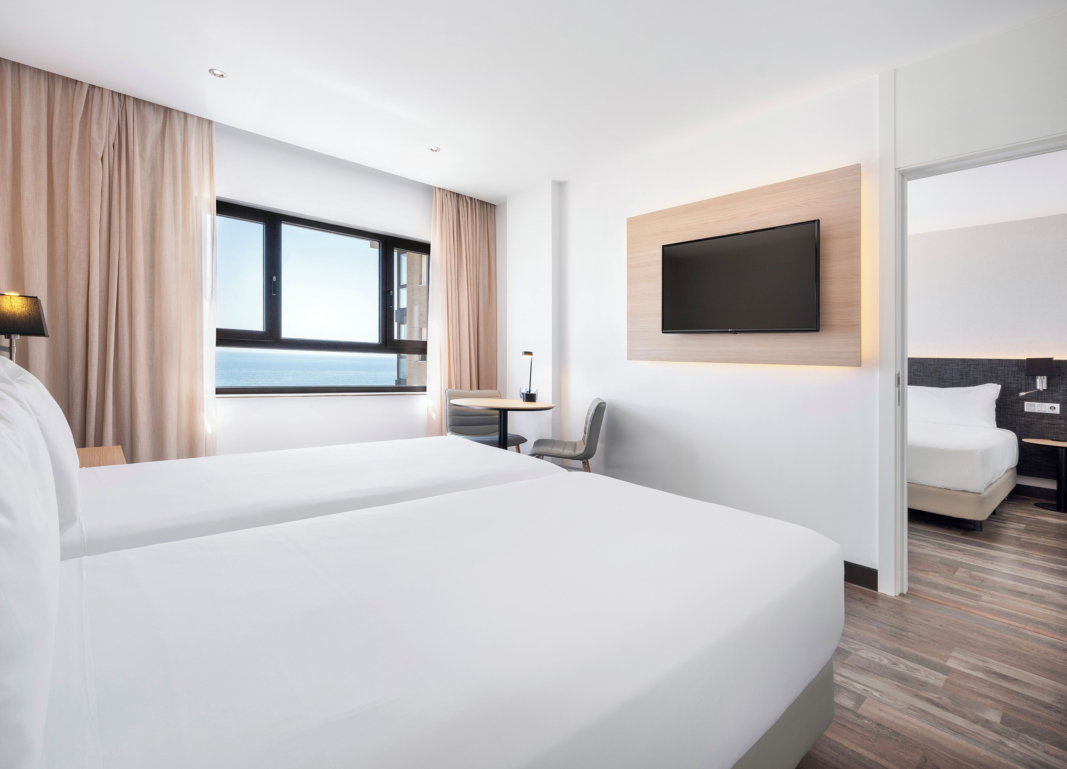A modern hotel room with two beds, a large window with a view, a flat-screen TV mounted on the wall, and a small desk with a chair.