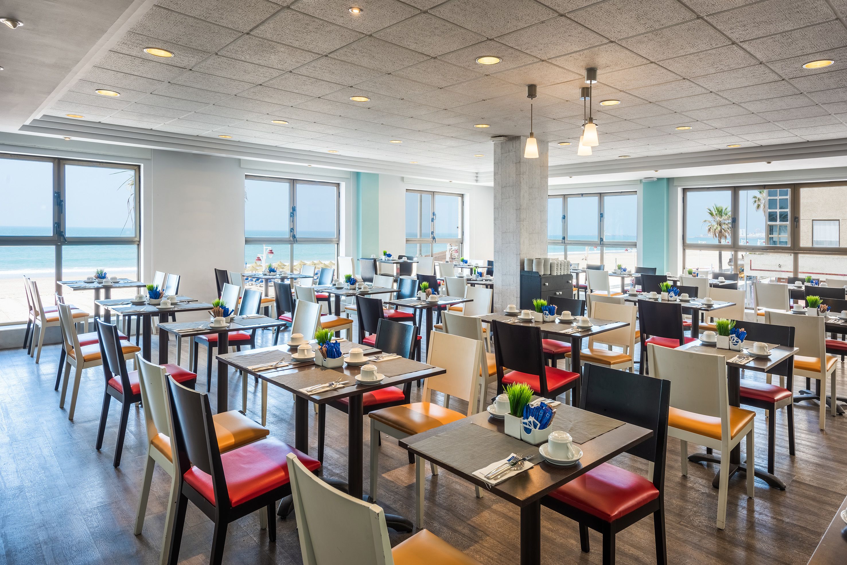 A spacious dining area with multiple tables and chairs, large windows offering a view of the ocean, and a modern ceiling design.