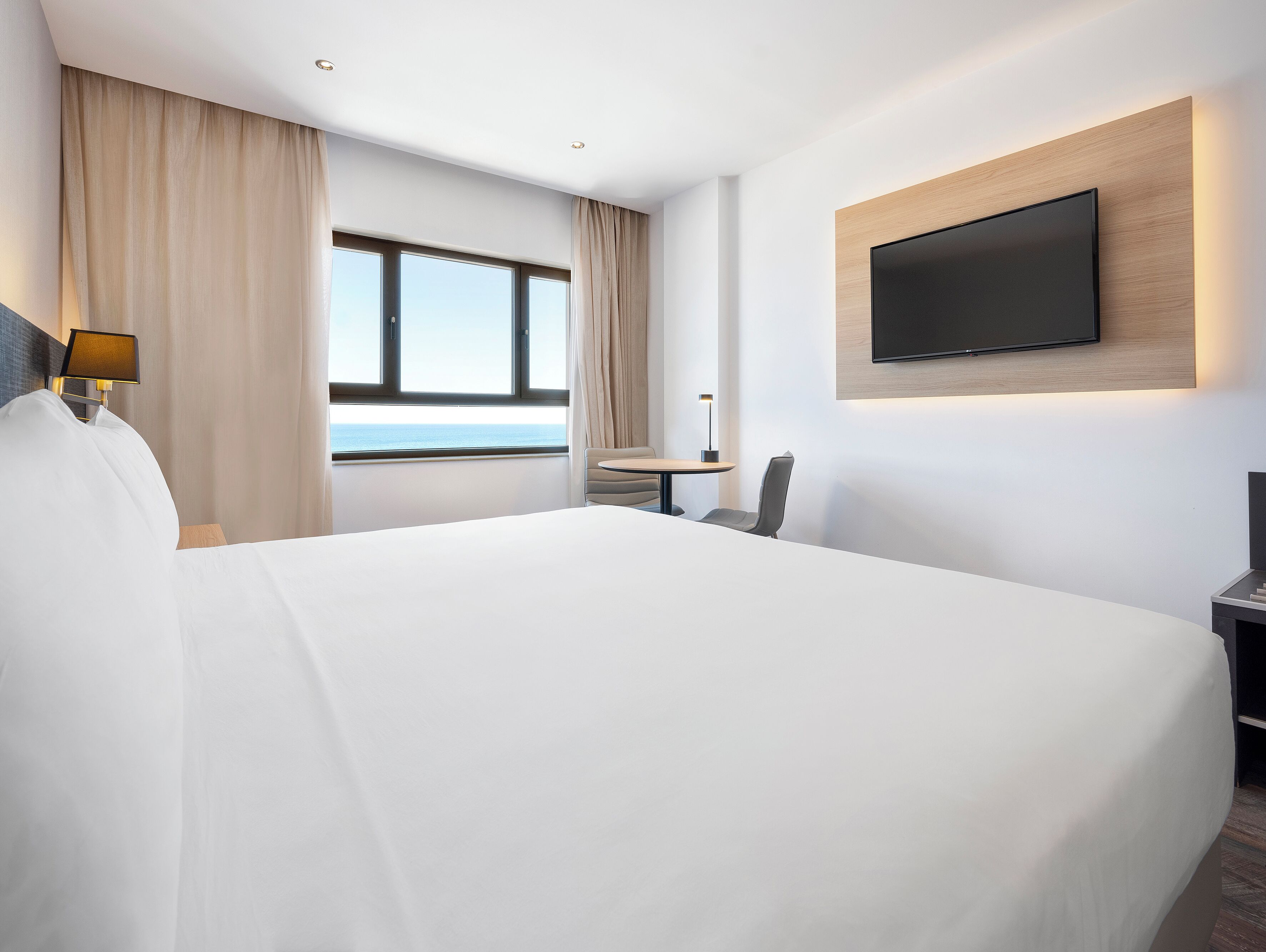 A modern hotel room with a large bed, a desk with a chair, a flat-screen TV mounted on the wall, and a window with a view of the sea.