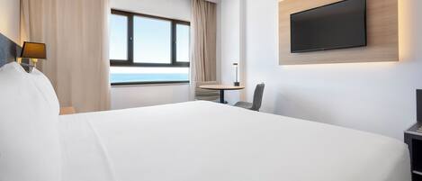 A modern hotel room with a large bed, a desk with a chair, a flat-screen TV mounted on the wall, and a window with a view of the sea.