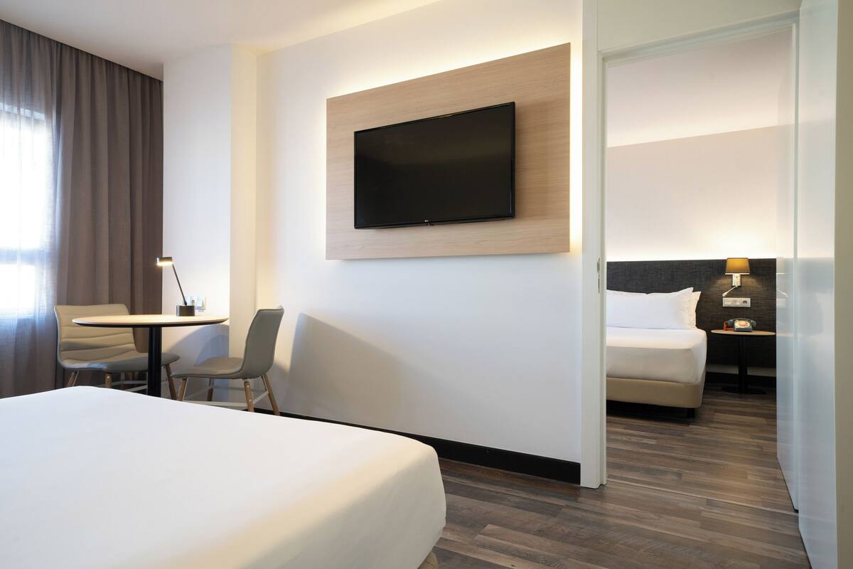 A hotel room with a large bed, a desk with two chairs, a flat-screen TV mounted on the wall, and a separate bedroom with a bed and nightstand.