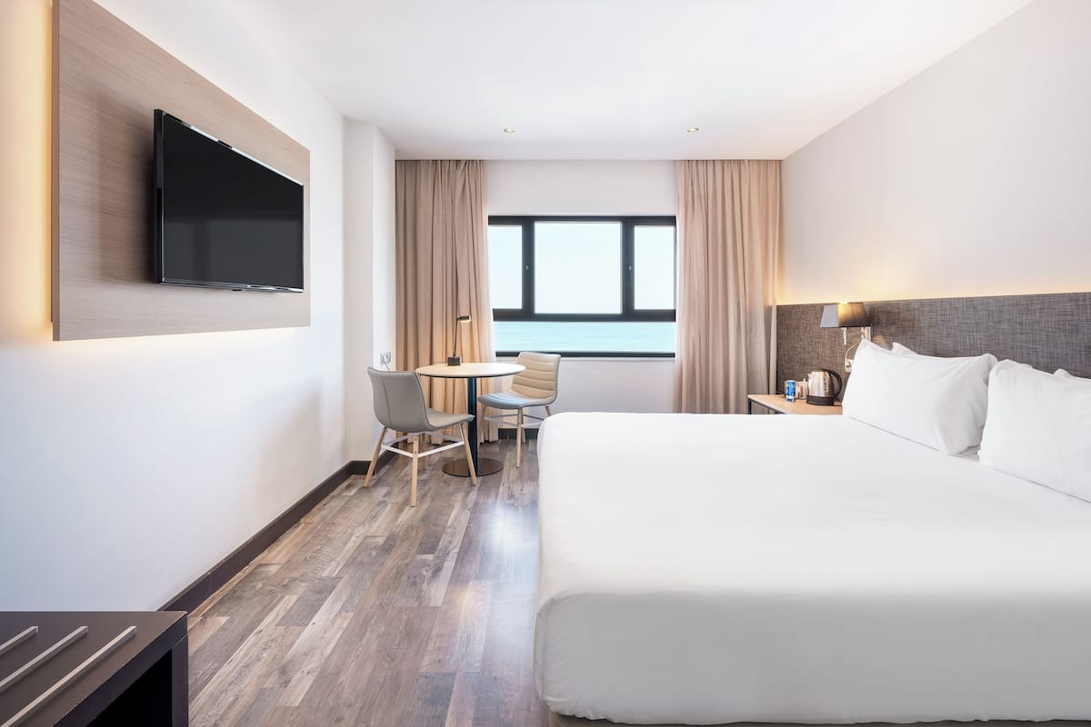 A hotel room with a large bed, a desk with a chair, a television mounted on the wall, and a window with curtains.