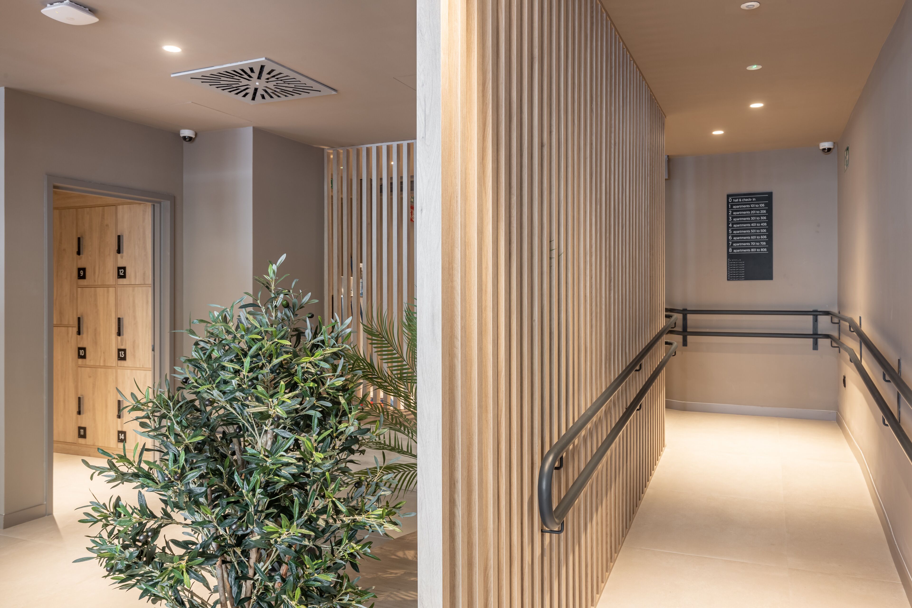 Un pasillo moderno de hotel con una puerta de madera, una planta, un panel de madera vertical y un letrero con números de habitaciones.