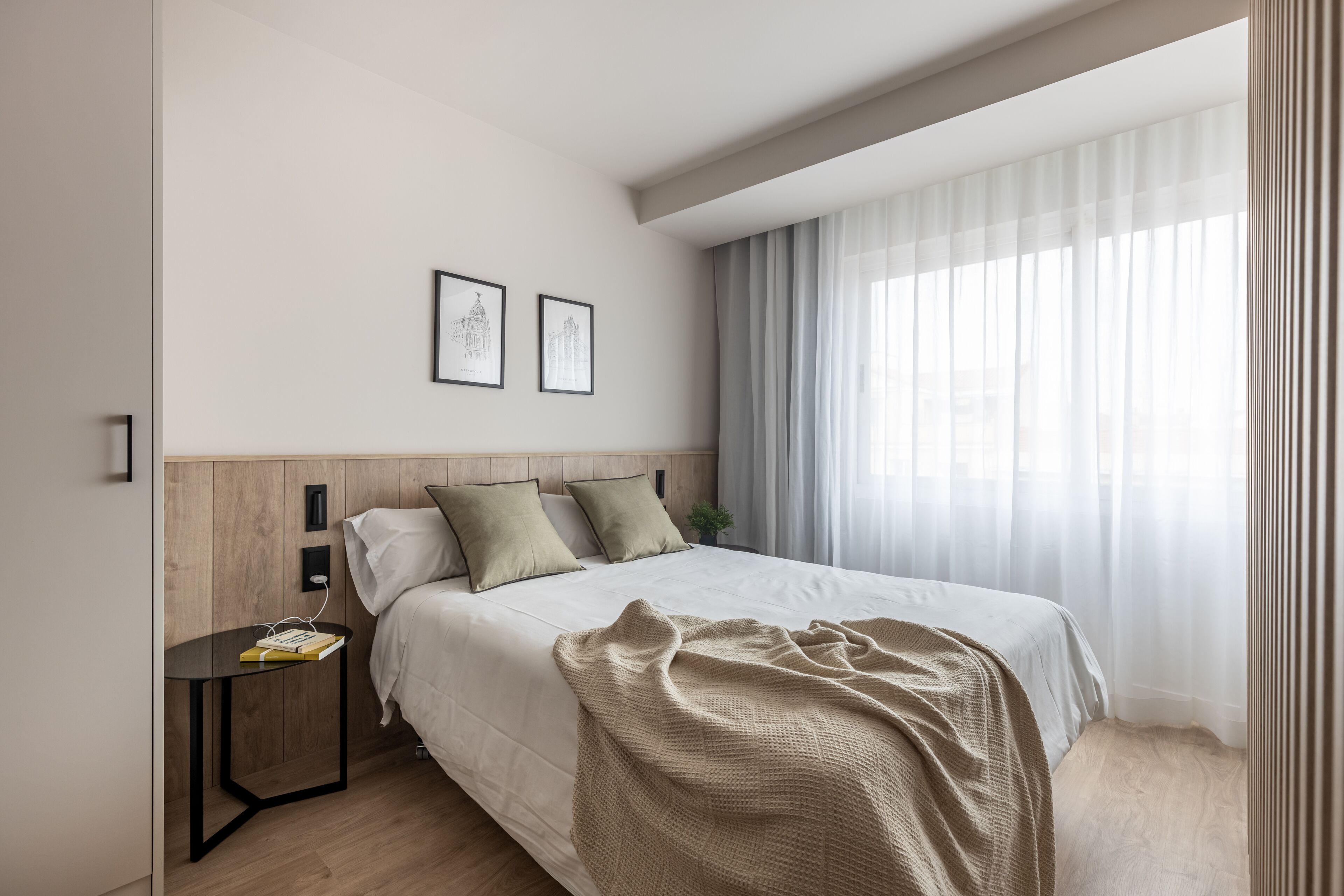 A modern bedroom with a bed, a nightstand, a window with sheer curtains, and two framed pictures on the wall.