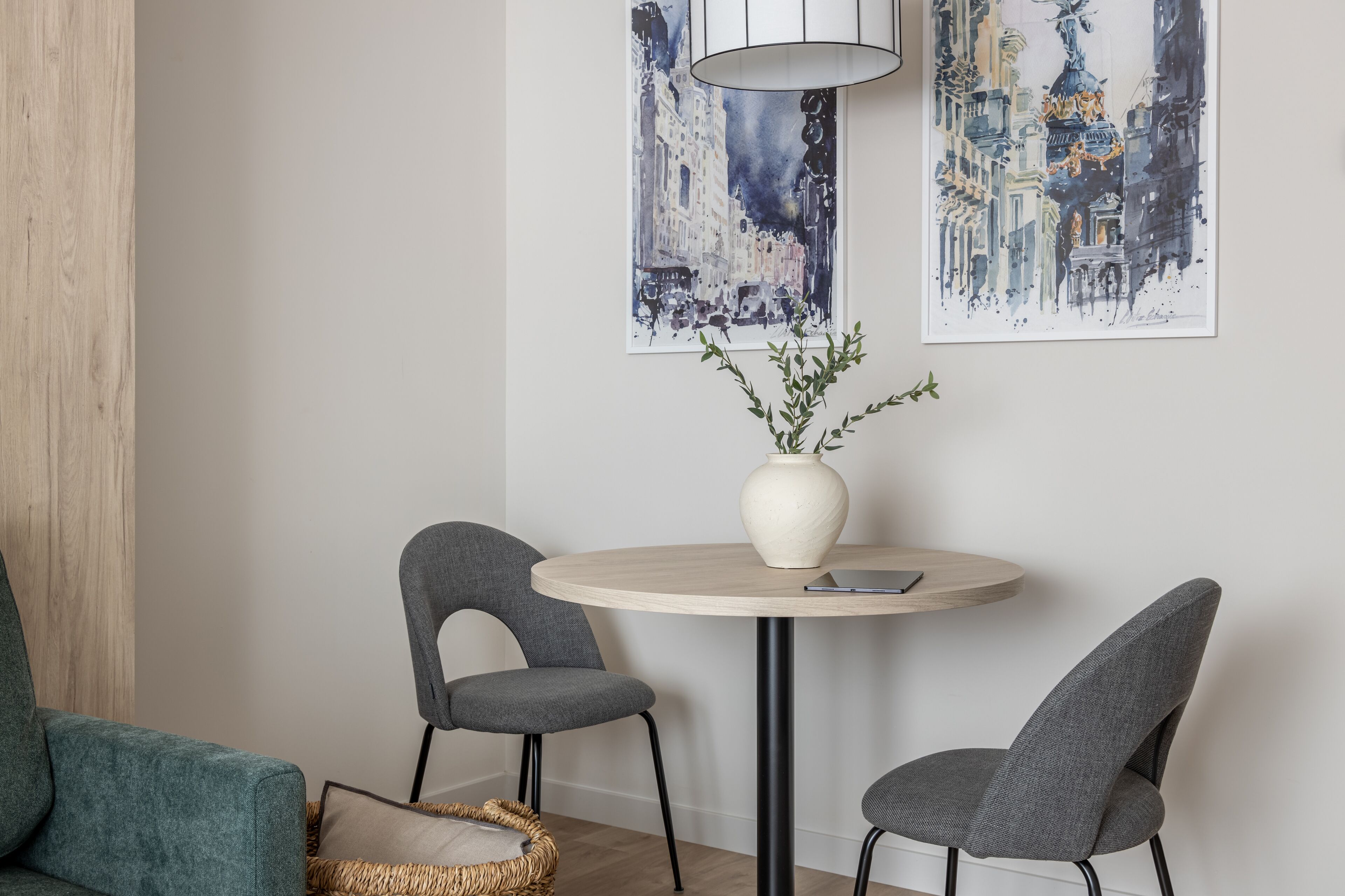 A modern room with a round table, two grey chairs, a white vase with greenery, and two abstract paintings on the wall.