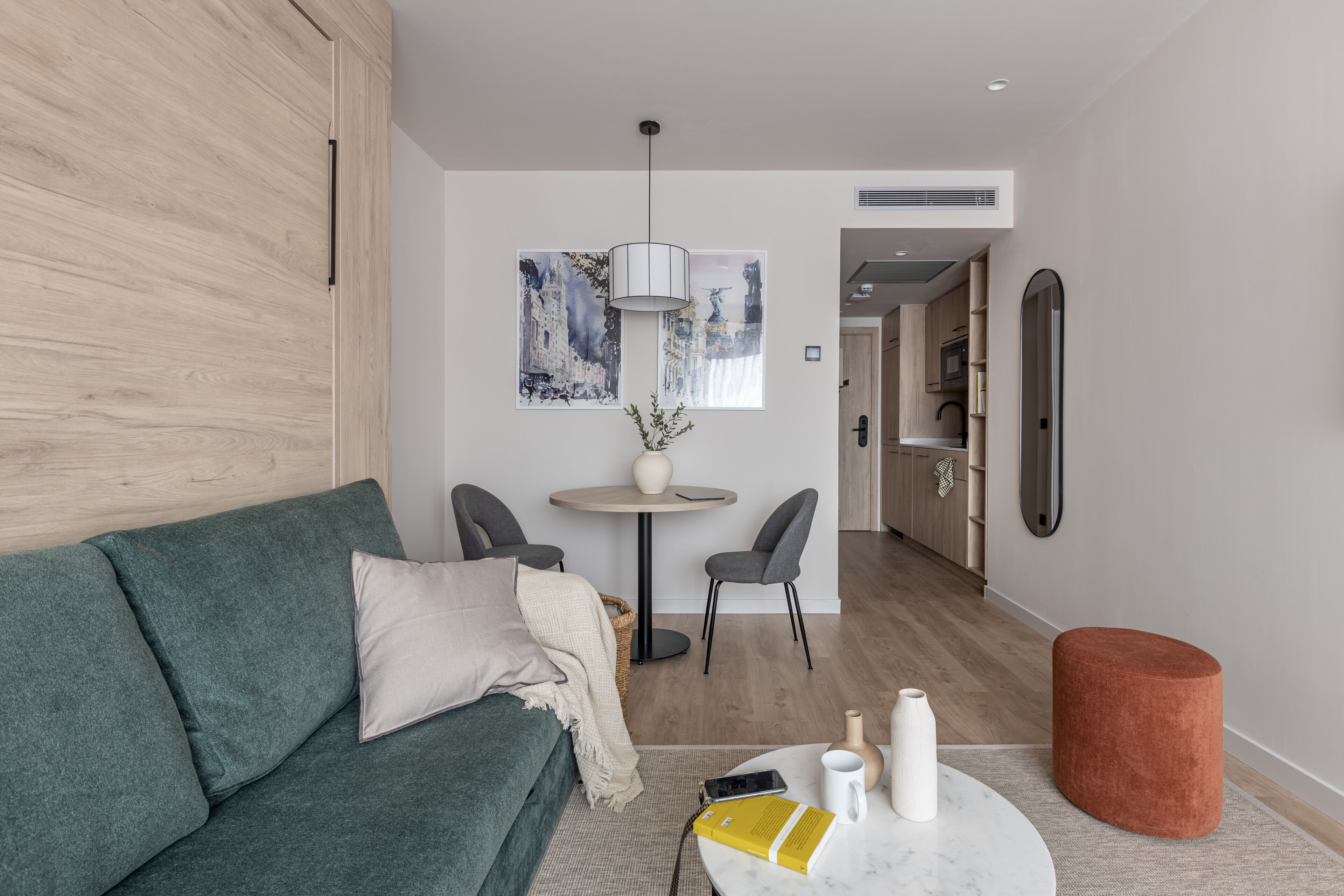 Un salon moderne avec un canapé vert, une table basse ronde et un petit pouf orange. On y trouve également un coin repas avec une table ronde et des chaises, ainsi qu’une cuisine en arrière-plan.