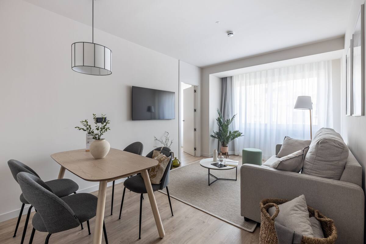 A modern living room with a sofa, a dining table with chairs, a TV mounted on the wall, and a large window with curtains.