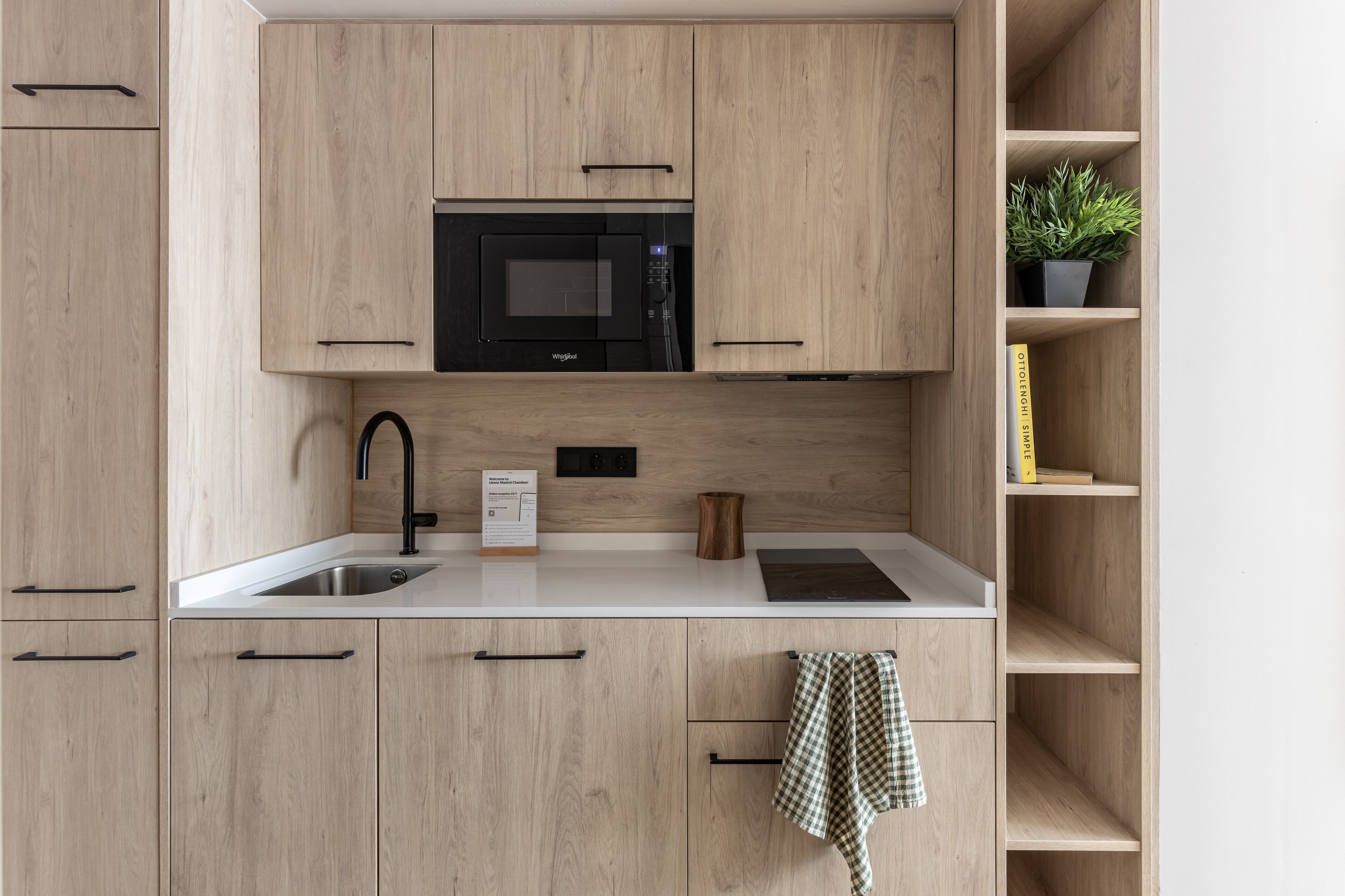 A modern kitchen with wooden cabinets, a black microwave, a sink, and a potted plant on a shelf.