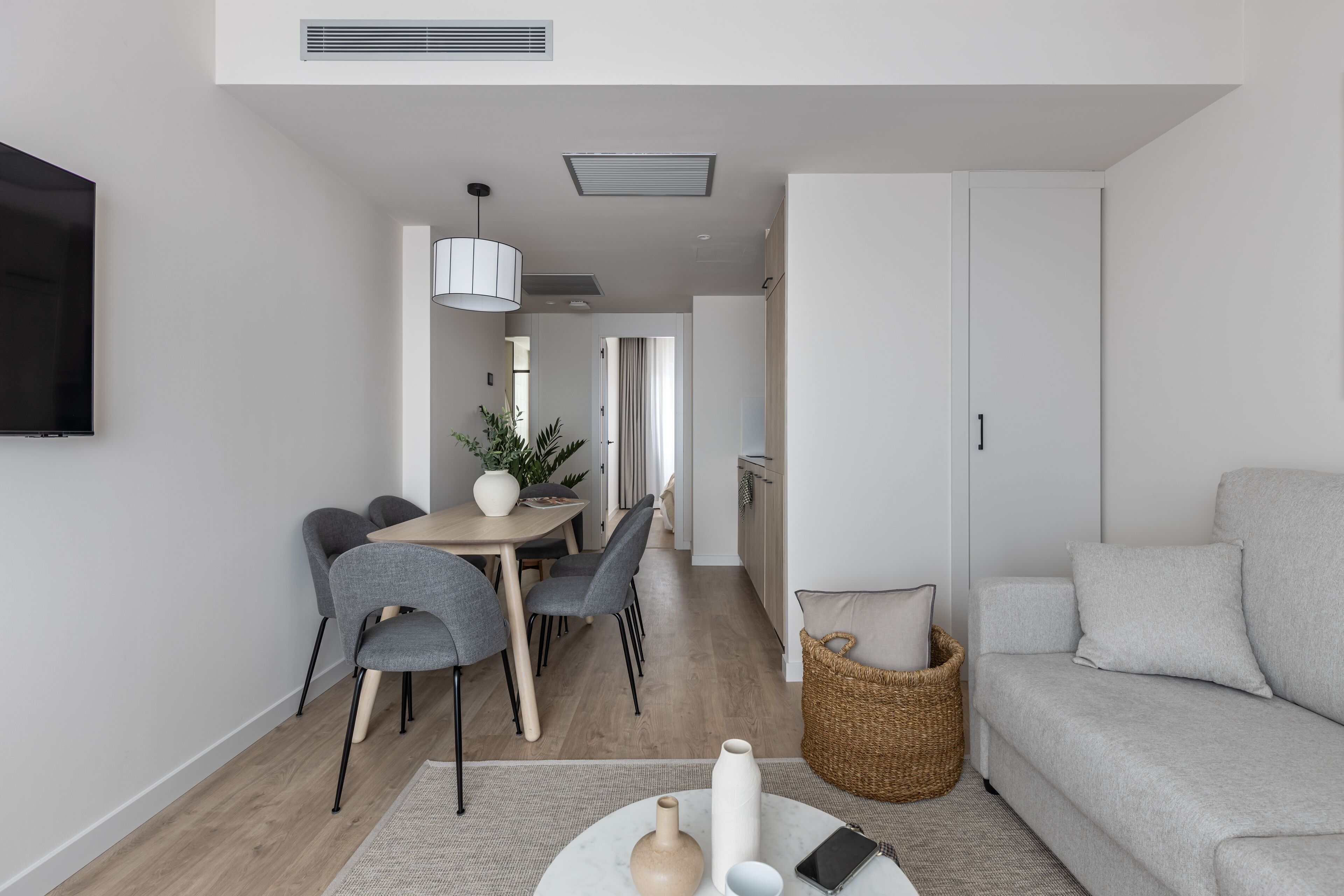 Un salon moderne avec un canapé gris, un coin repas avec une table et des chaises, et une cuisine en arrière-plan.