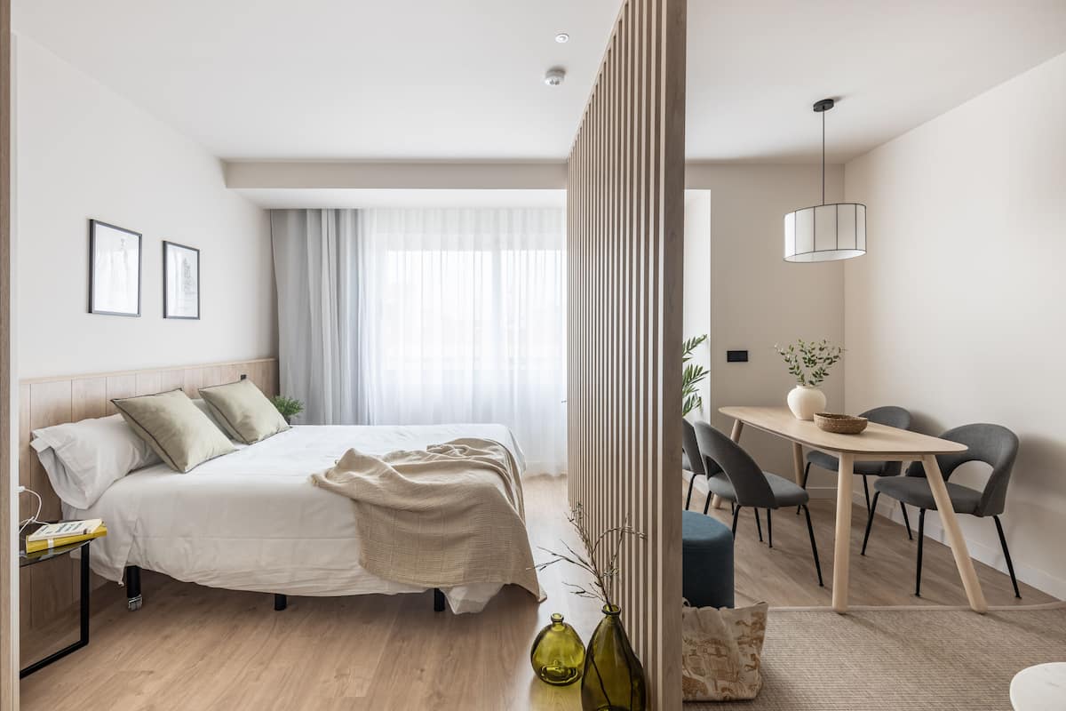 A modern hotel room with a large bed, bedside tables, a dining area with chairs, and a view of the outdoors.