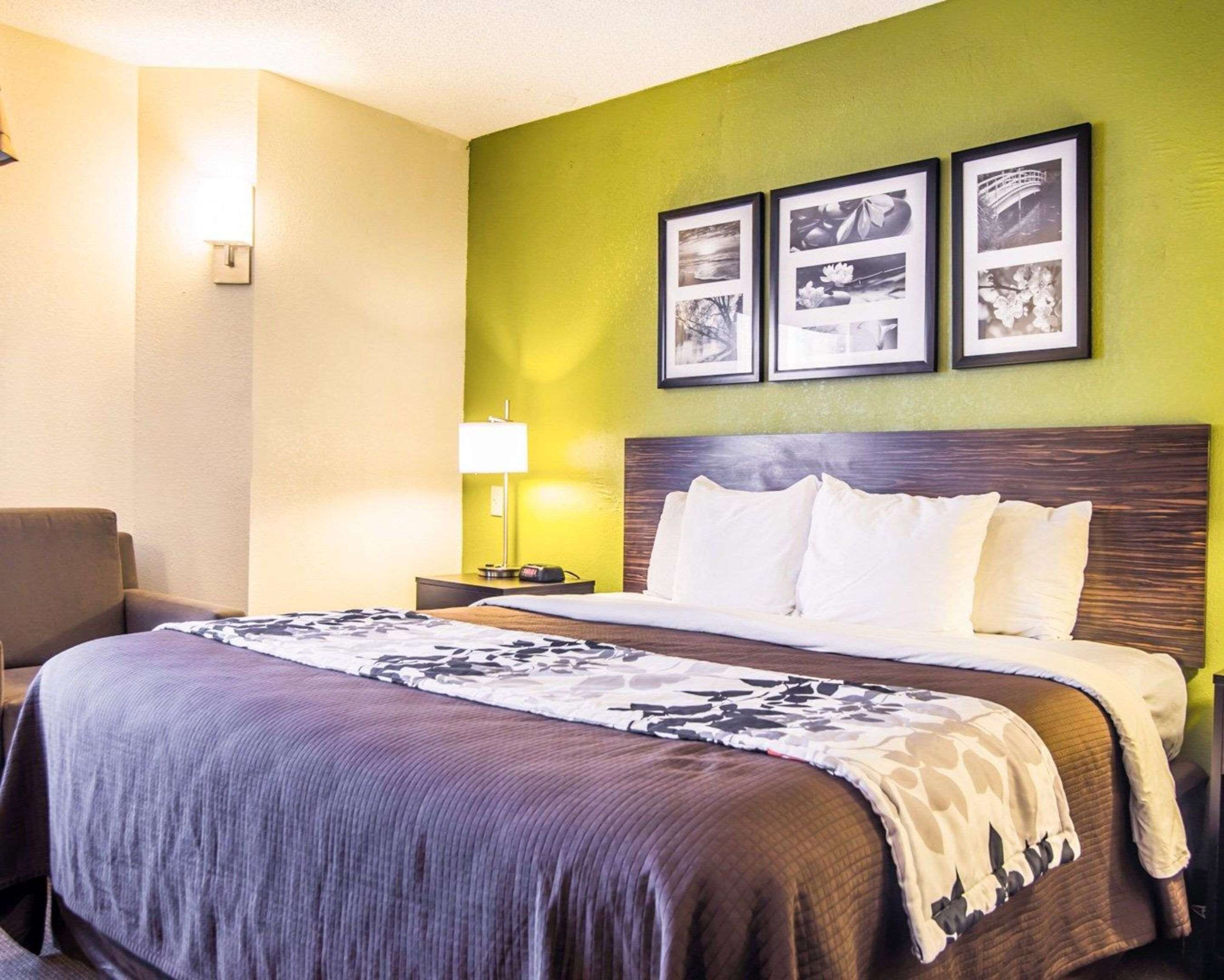 Ein Hotelzimmer mit einem Bett, einem Nachttisch, einer Lampe und einem Sessel. Über dem Bett hängen drei gerahmte Bilder.