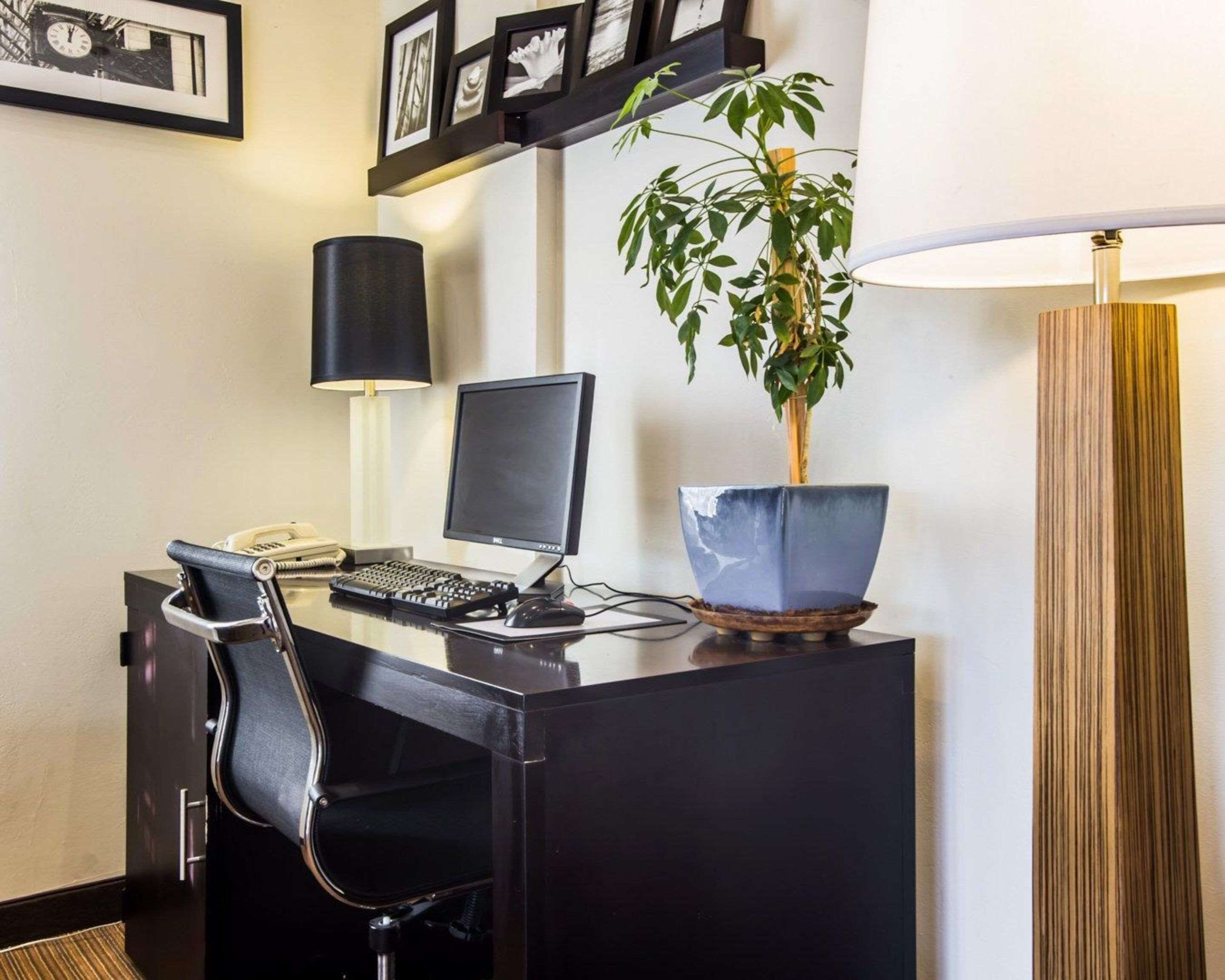 Habitación de hotel con escritorio, computadora, silla, lámpara, planta en maceta y lámpara de pie.