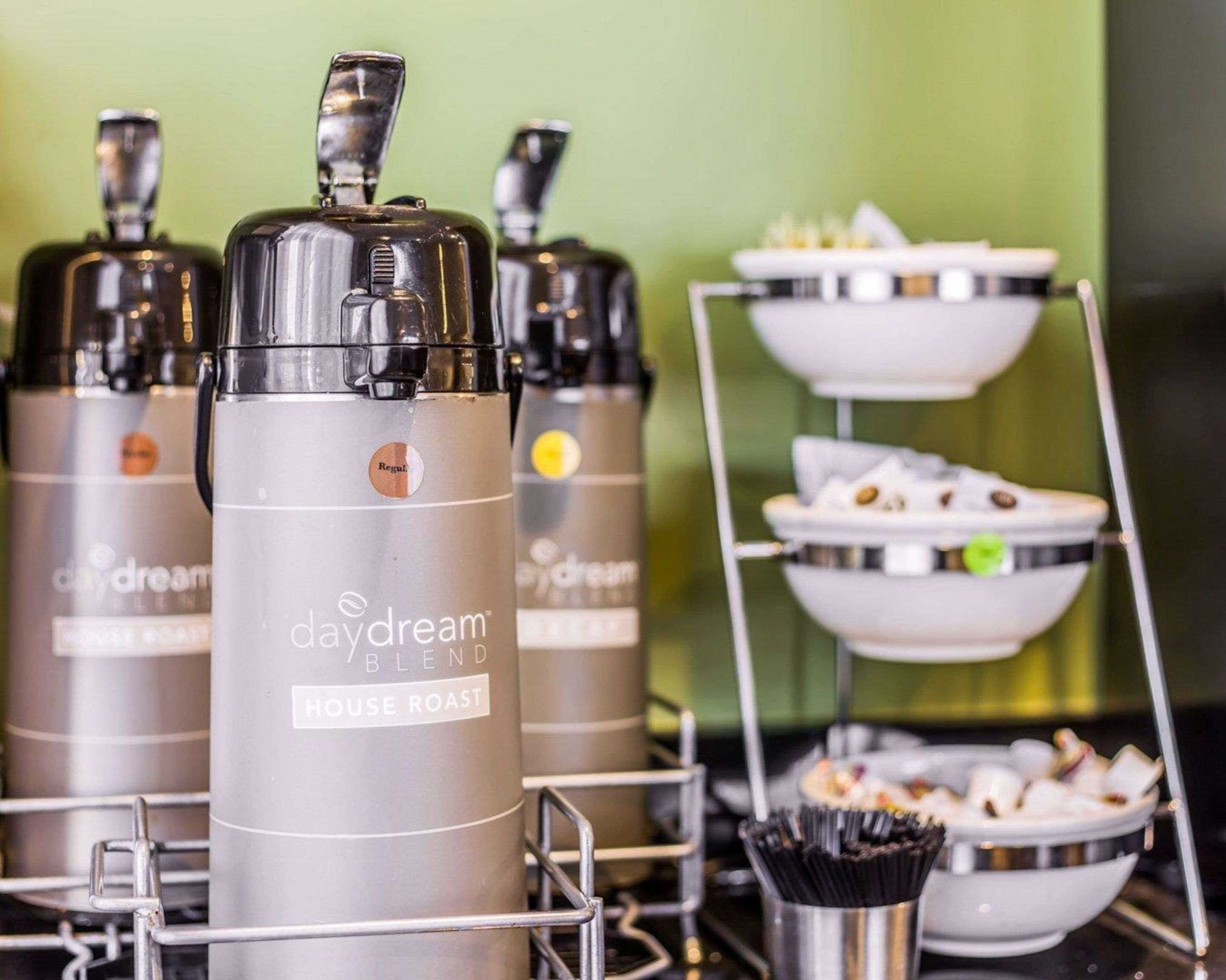 A coffee station with multiple coffee dispensers labeled 'daydream' and 'house roast', a tiered tray with pastries, and a metal basket with coffee stirrers.