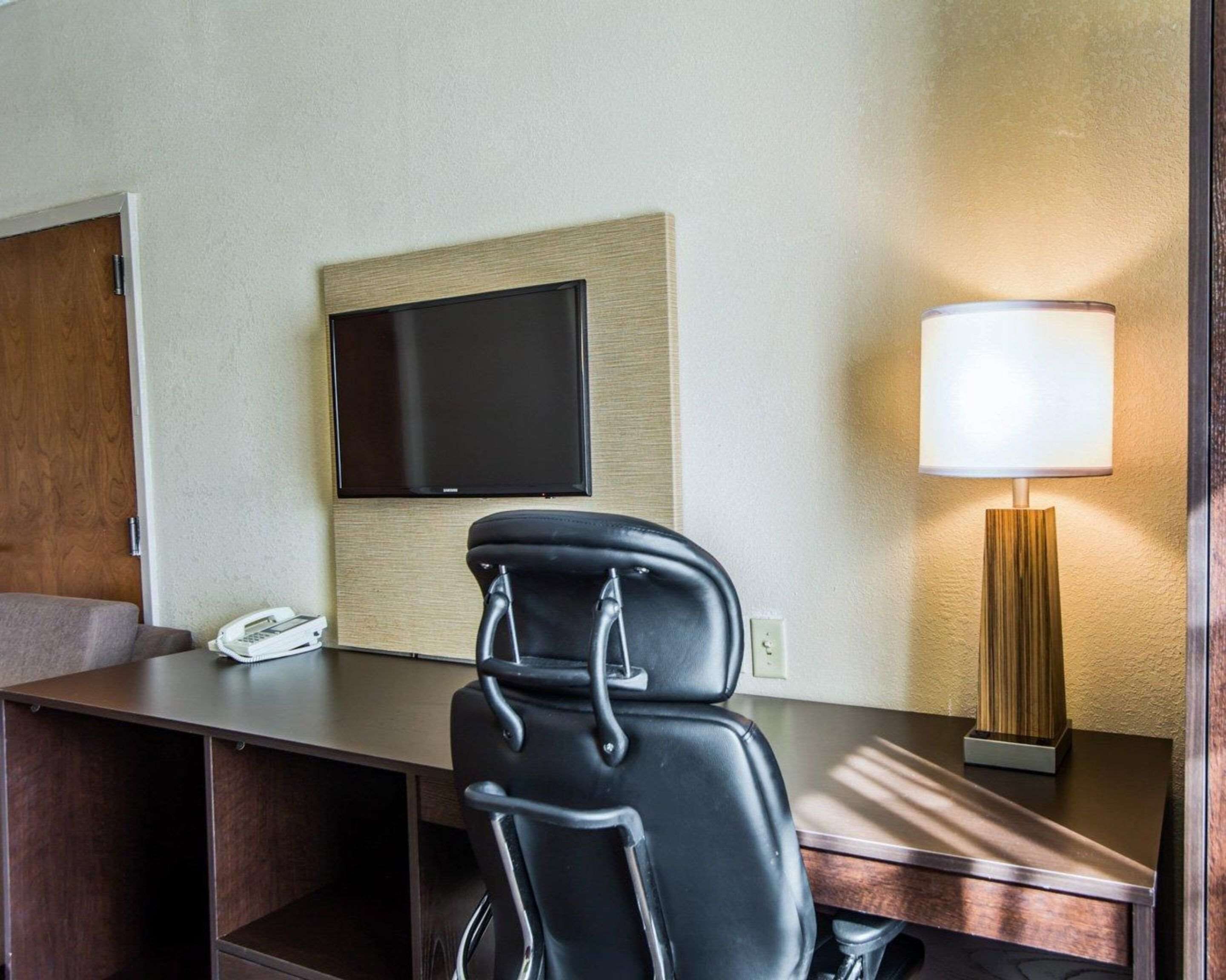 A hotel room with a desk, a chair, a flat-screen TV mounted on the wall, a bedside lamp, and a telephone.
