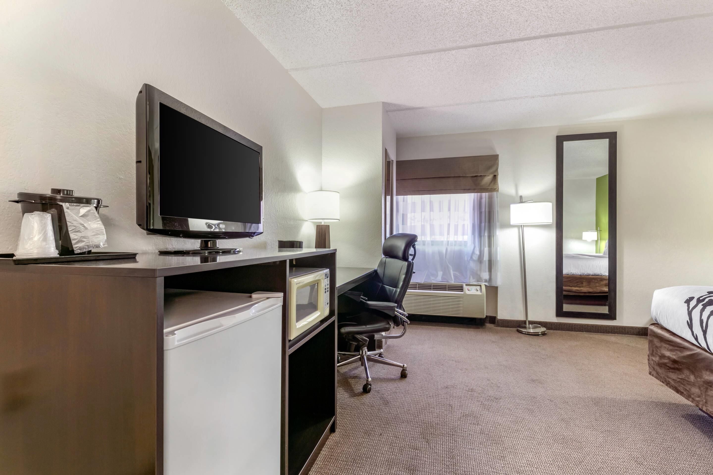 A hotel room with a flat-screen TV, a desk with a microwave, a chair, a bed, a large mirror, and a window with curtains.