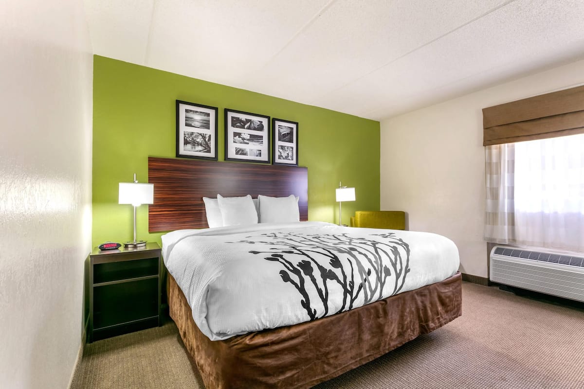 Une chambre d’hôtel comprenant un lit, des tables de chevet, une petite table, une lampe, un climatiseur fixé au mur et des tableaux encadrés accrochés au mur.