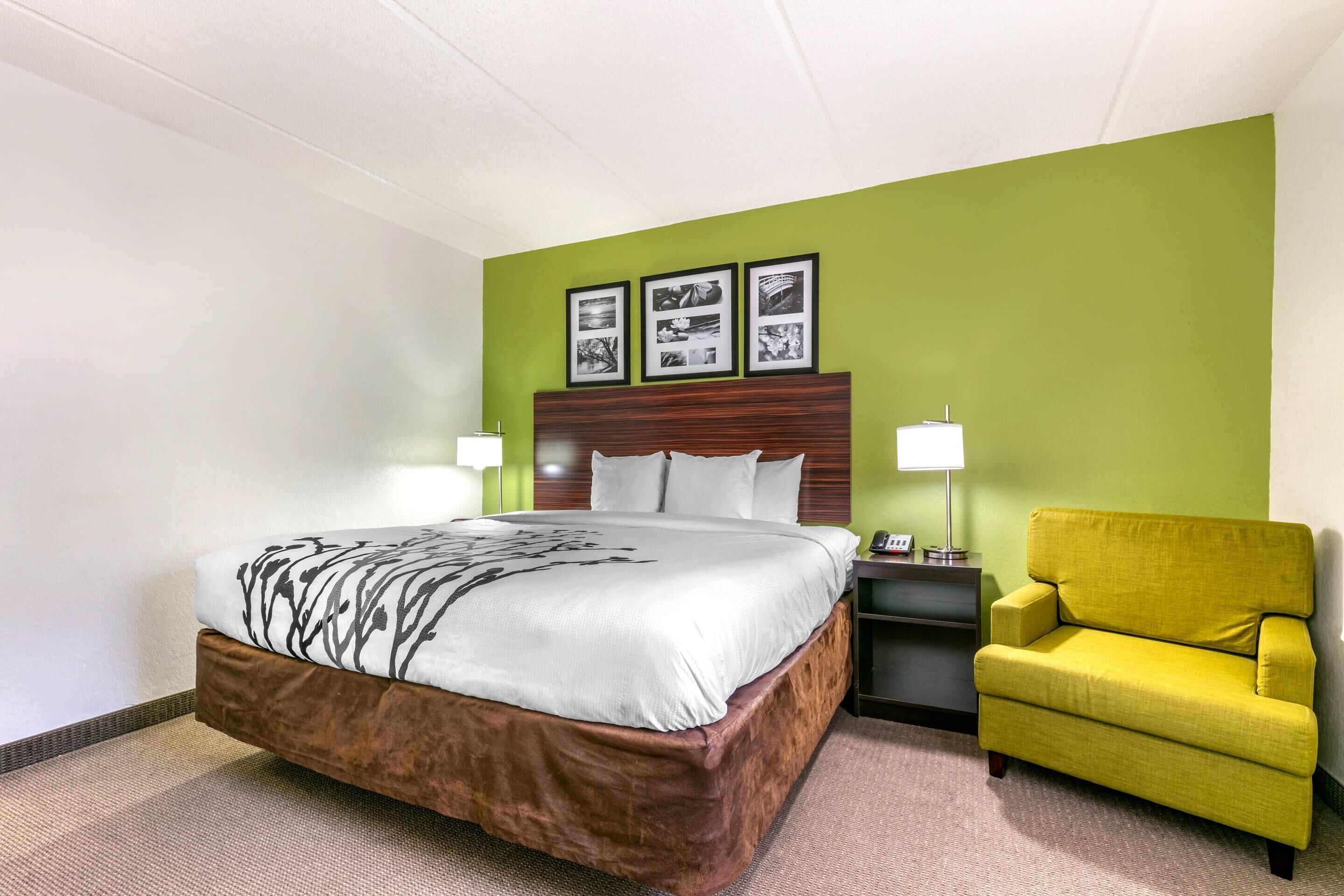 A hotel room with a large bed, a yellow armchair, a nightstand with a lamp, and three framed pictures on the wall.