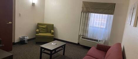 A hotel room with a sofa, a chair, a small table, a window with curtains, and a wall-mounted air conditioner.