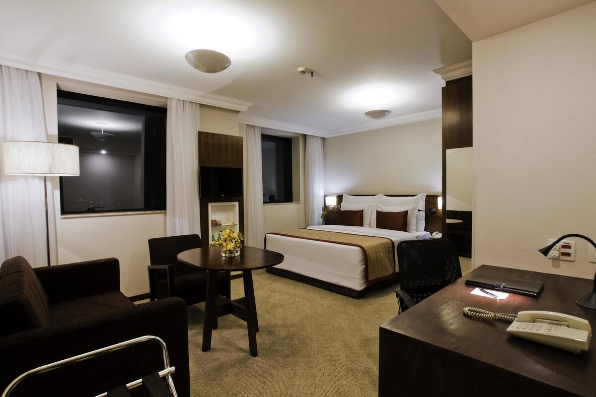 A hotel room with a bed, a desk with a telephone, a chair, a small table with a vase of flowers, and a television on the wall.