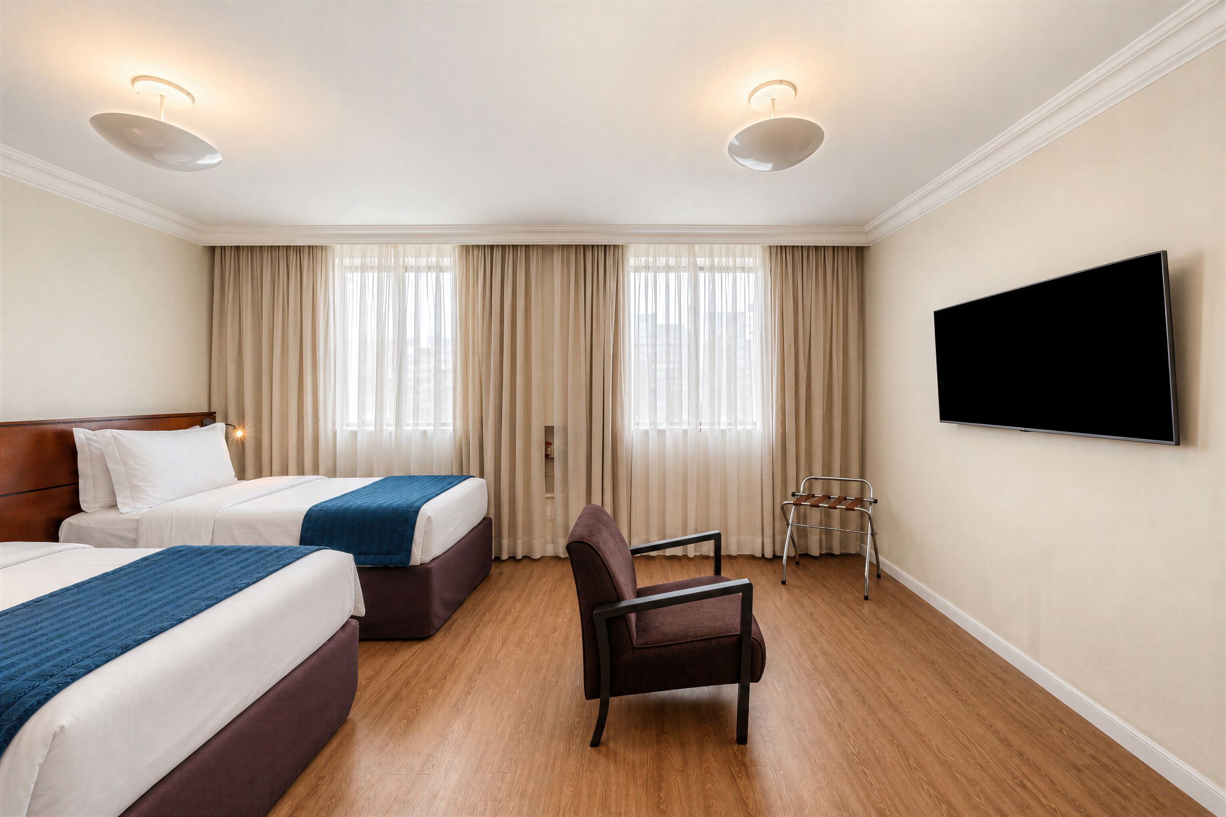 Ein Hotelzimmer mit zwei Betten, einem Fernseher, einem Sessel und einem Fenster mit Vorhängen.