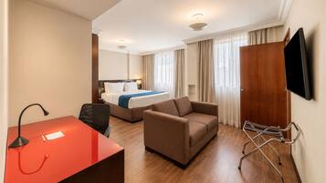Quarto de hotel com cama, escrivaninha, cadeira, sofá, televisão fixada na parede e janela com cortinas.