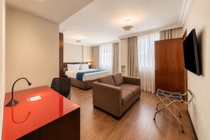 Habitación de hotel con cama, escritorio, silla, sofá, televisor montado en la pared y ventana con cortinas.