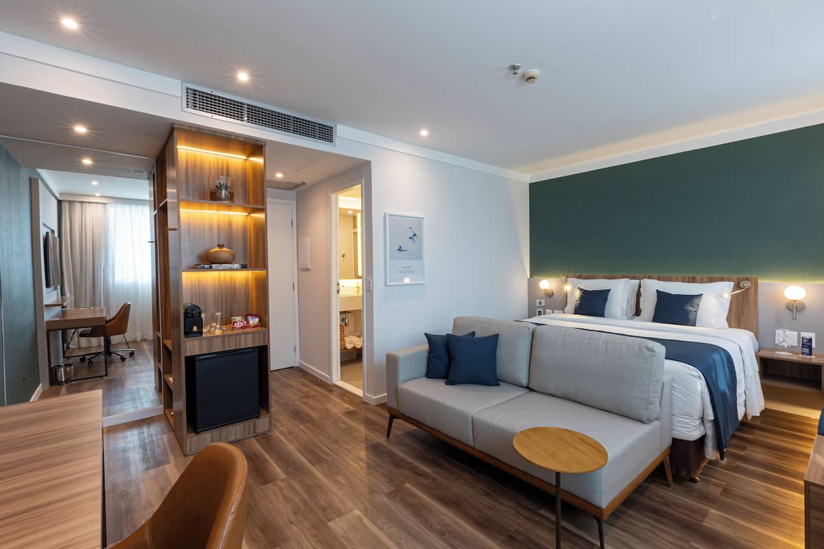 A modern hotel room with a sofa, a bed with blue bedding, a wooden desk, and a small table.