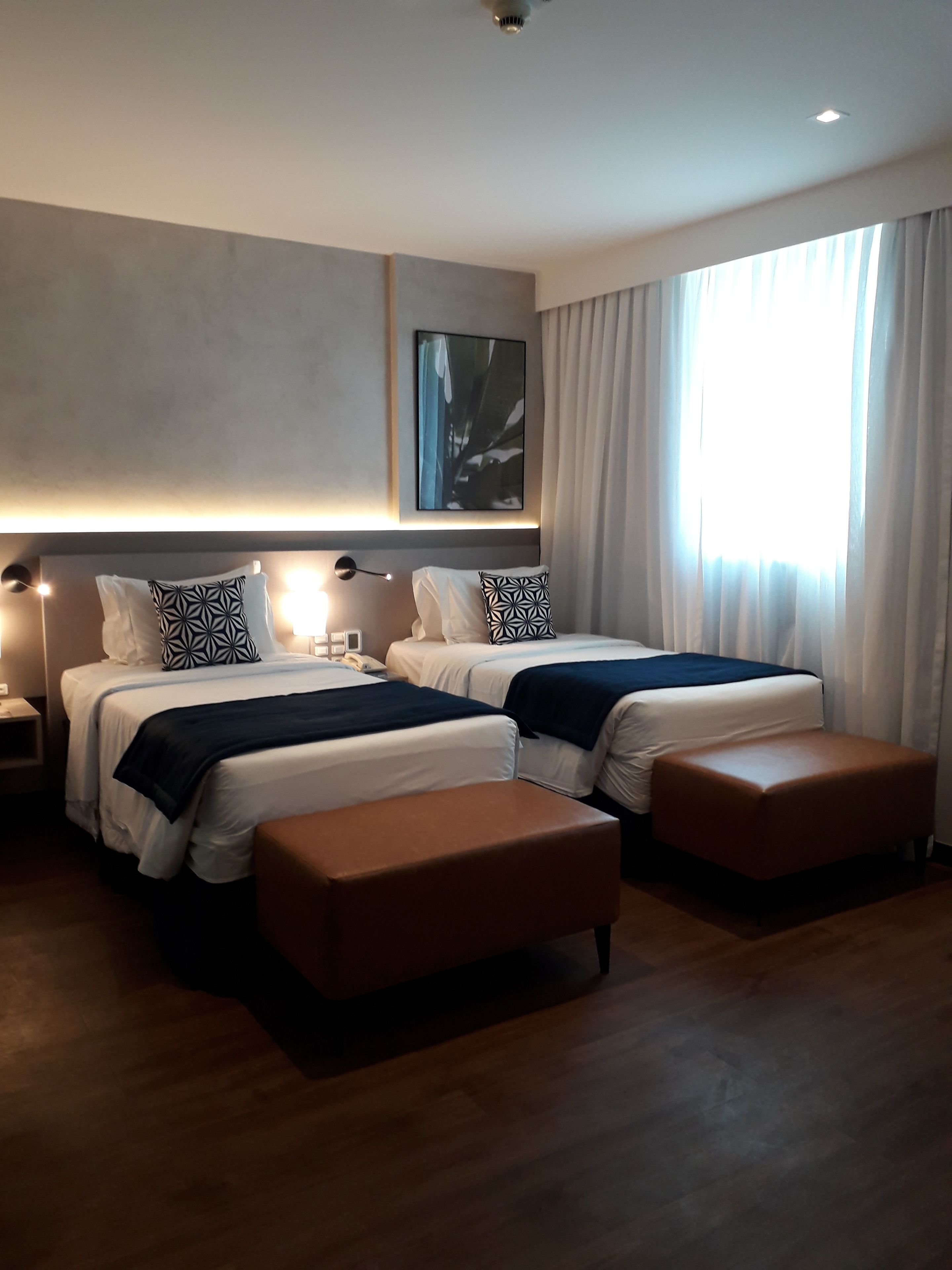 Ett modernt hotellrum med två sängar, ett stort fönster med gardiner, ett konstverk som hänger på väggen och ett nattduksbord med en lampa.