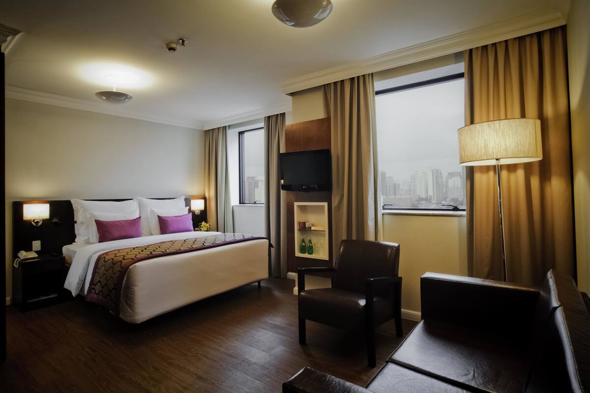 A hotel room with a large bed, a desk, a chair, a TV, and a window with a city view.
