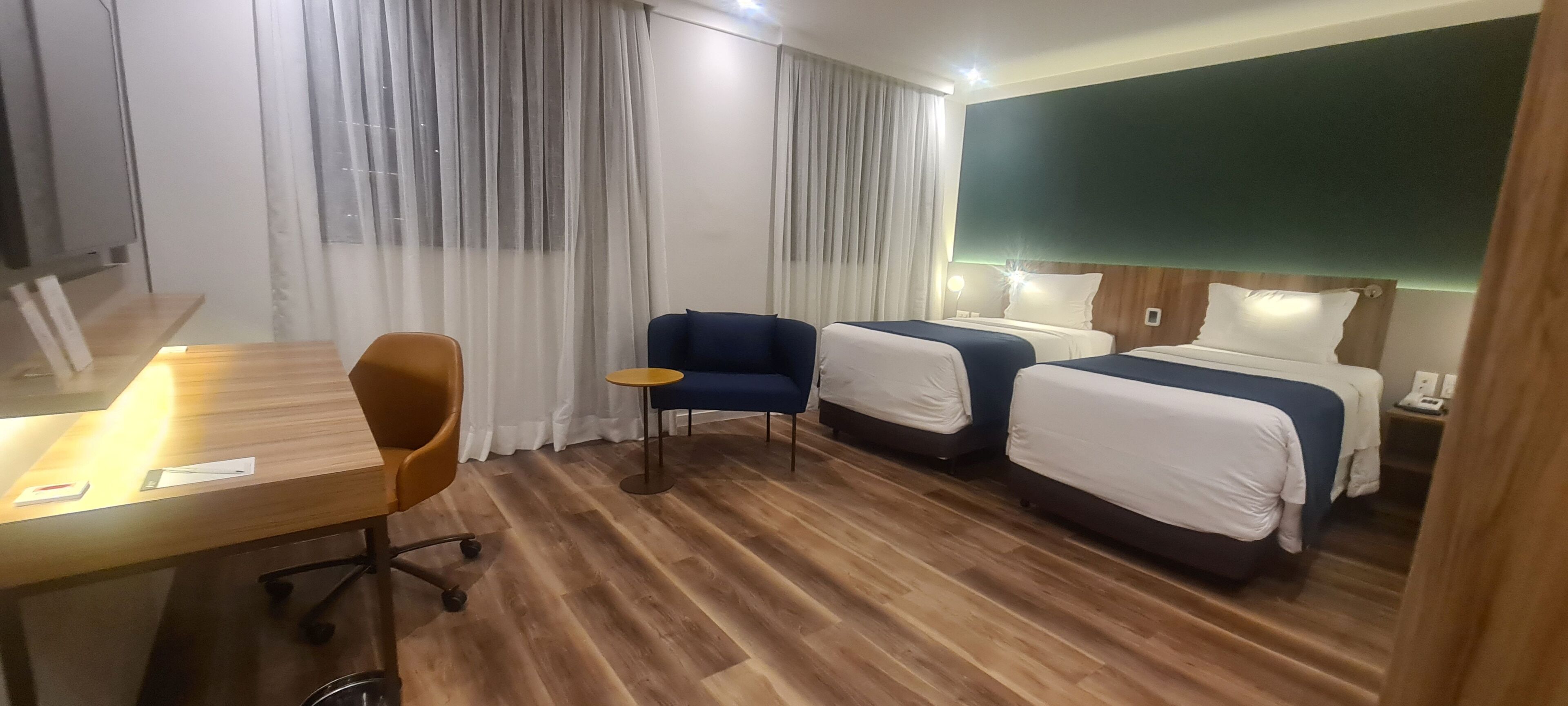 A hotel room with a wooden floor, two beds with white and blue bedding, a desk with a chair, and a small round table.