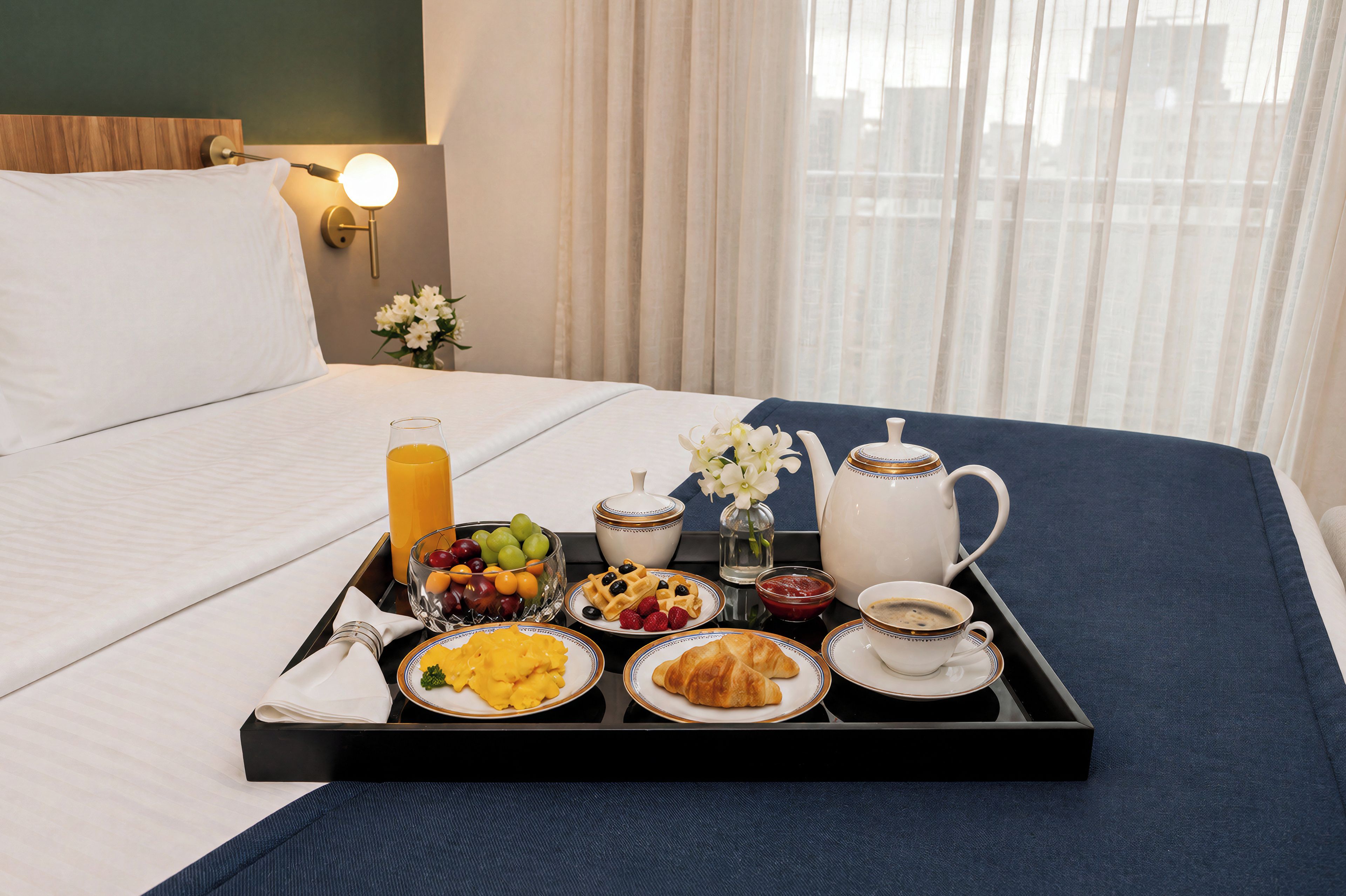 A neatly arranged breakfast tray on a bed with a view of a cityscape through sheer curtains.