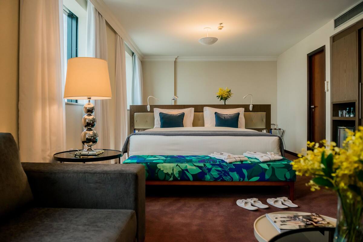 A hotel room with a bed, a bedside table with a lamp, a chair, a small table with a magazine, and a vase with flowers.