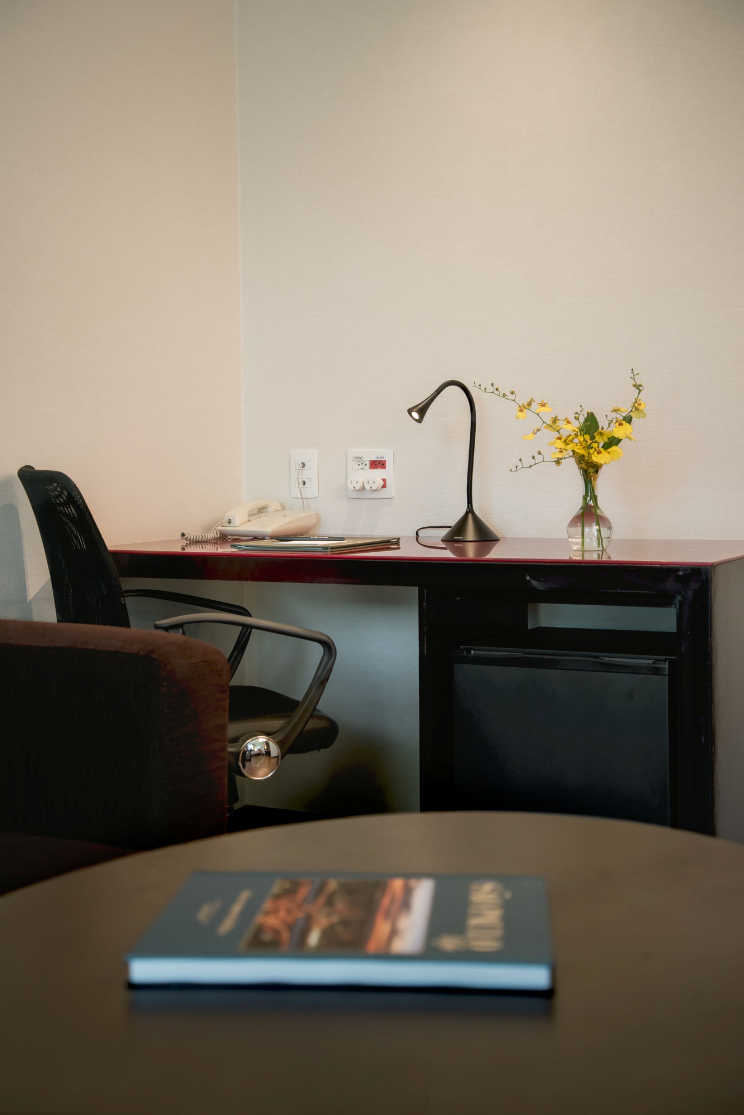 A hotel room with a desk, a chair, a lamp, a vase with flowers, and a book on the table.