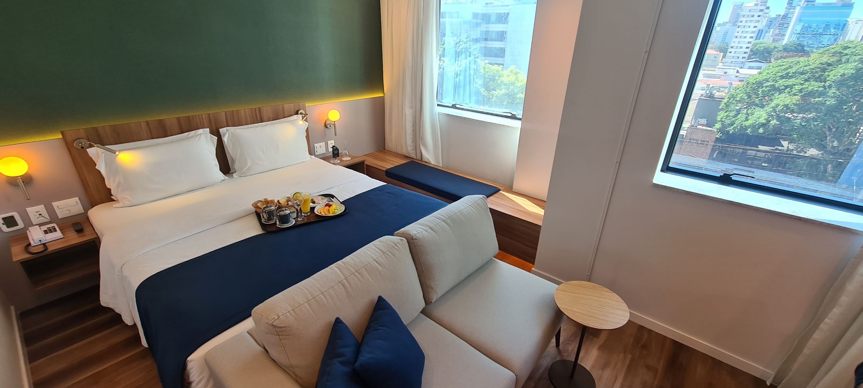 A hotel room with a bed, a sofa, a small table, and a window with a city view.