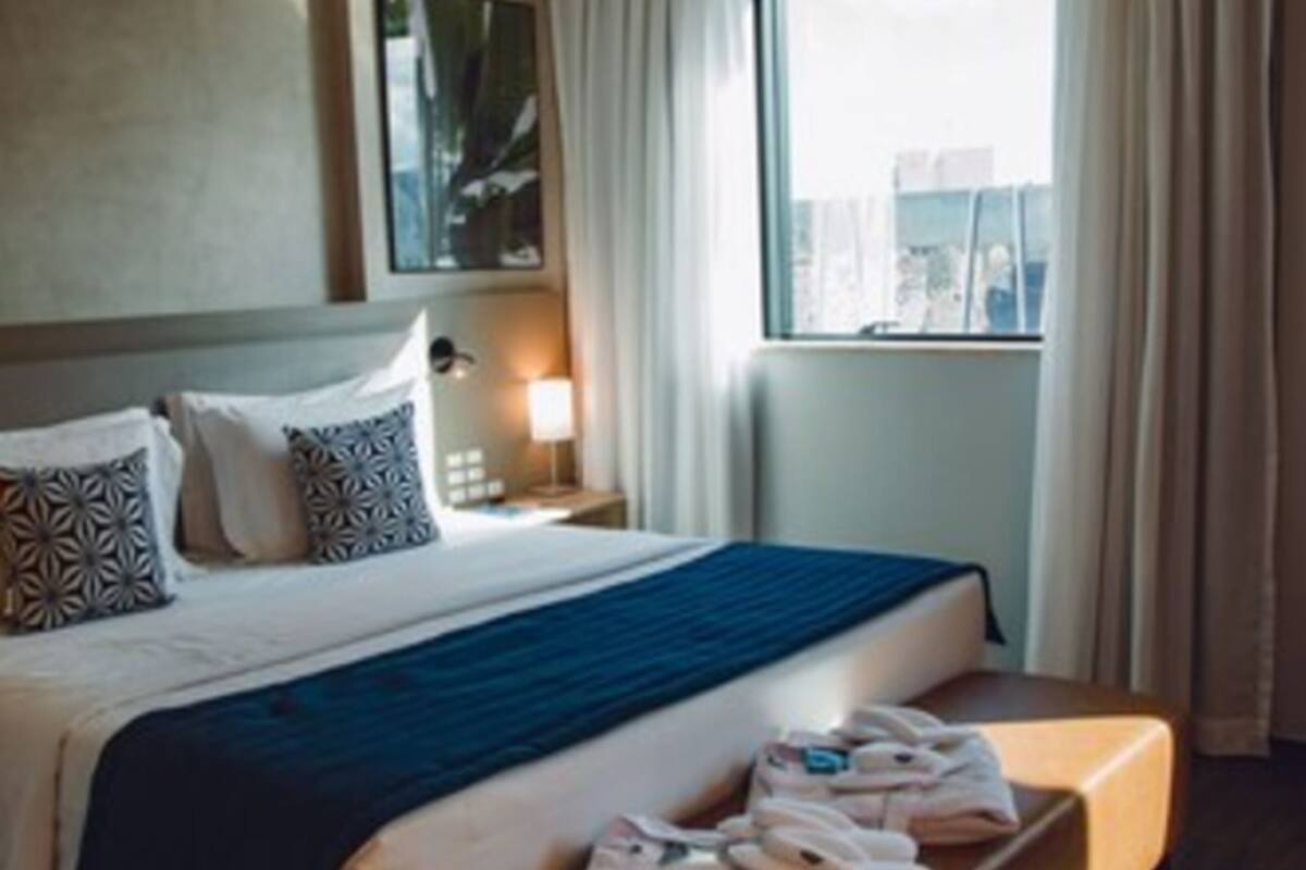 A hotel room with a large bed, a bedside table with a lamp, a window with curtains, and a view of the city.