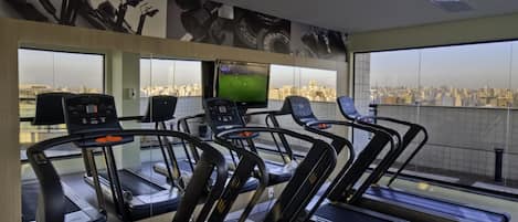 Un gimnasio con cintas de correr, televisión y vistas a la ciudad.