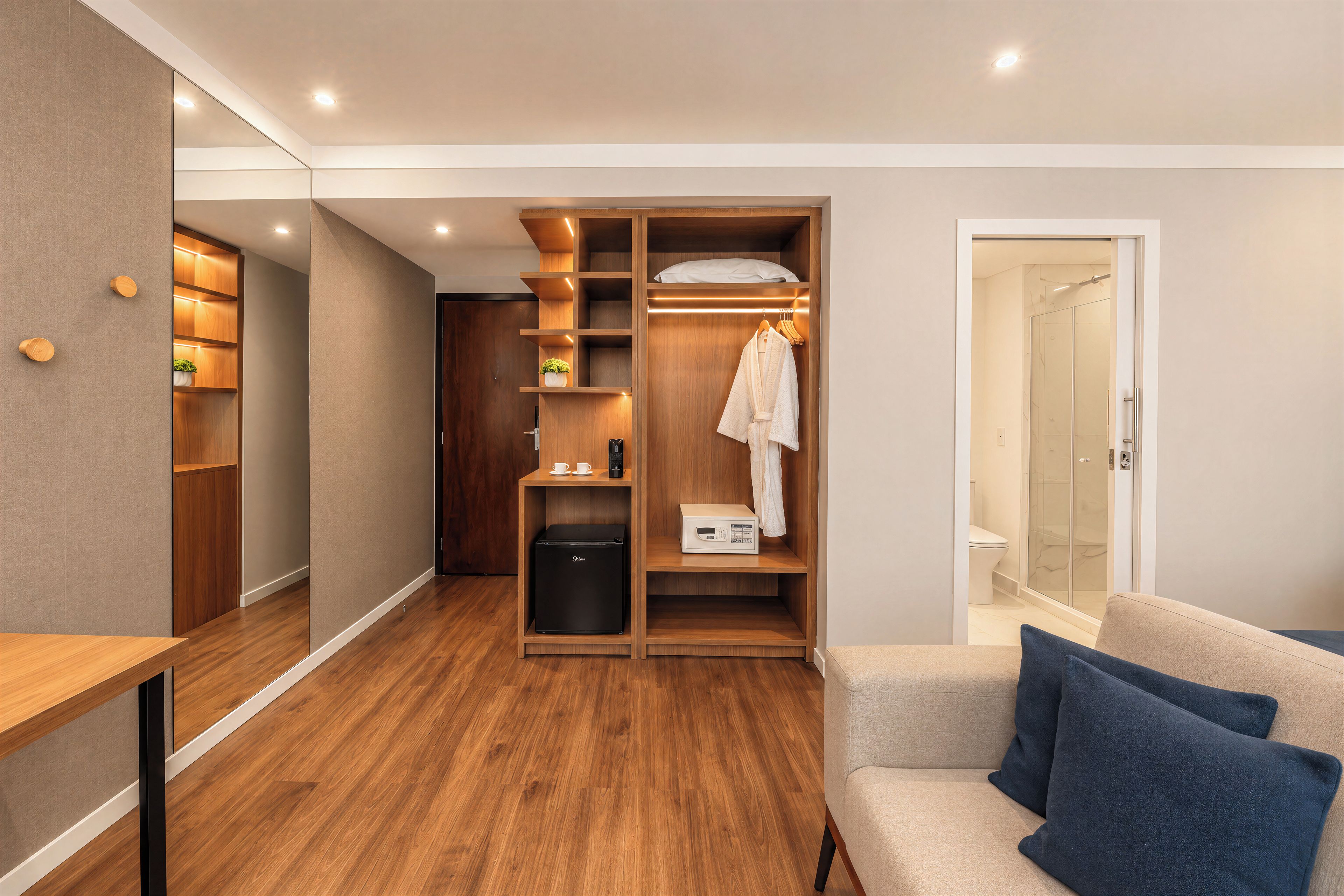 A compact hotel room with a walk-in closet, a small kitchenette, a shower area, and a seating area with cushions.