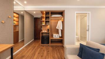 A compact hotel room with a walk-in closet, a small kitchenette, a shower area, and a seating area with cushions.
