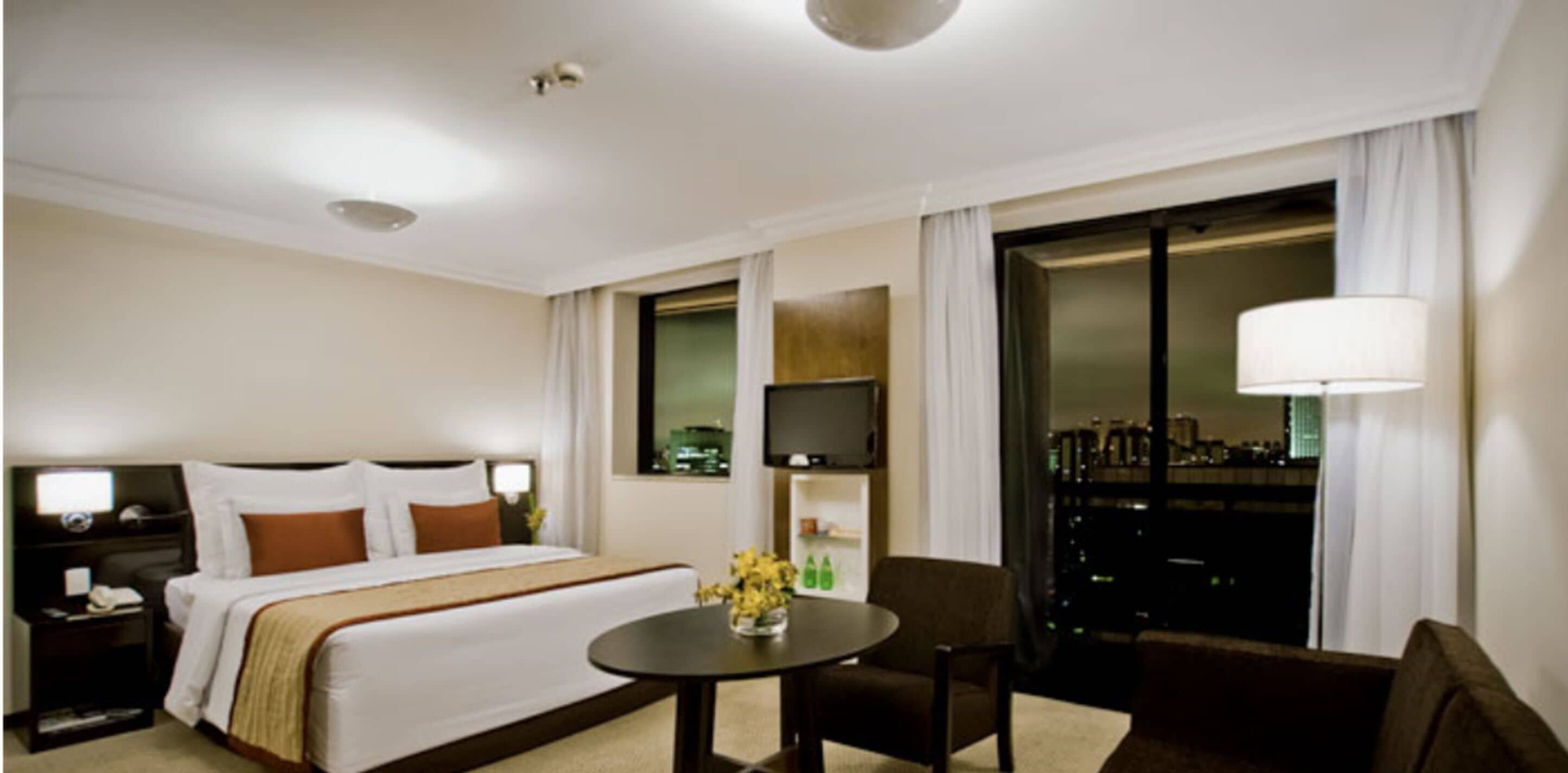 A hotel room with a large bed, a desk with a lamp, a chair, and a view of the city at night.