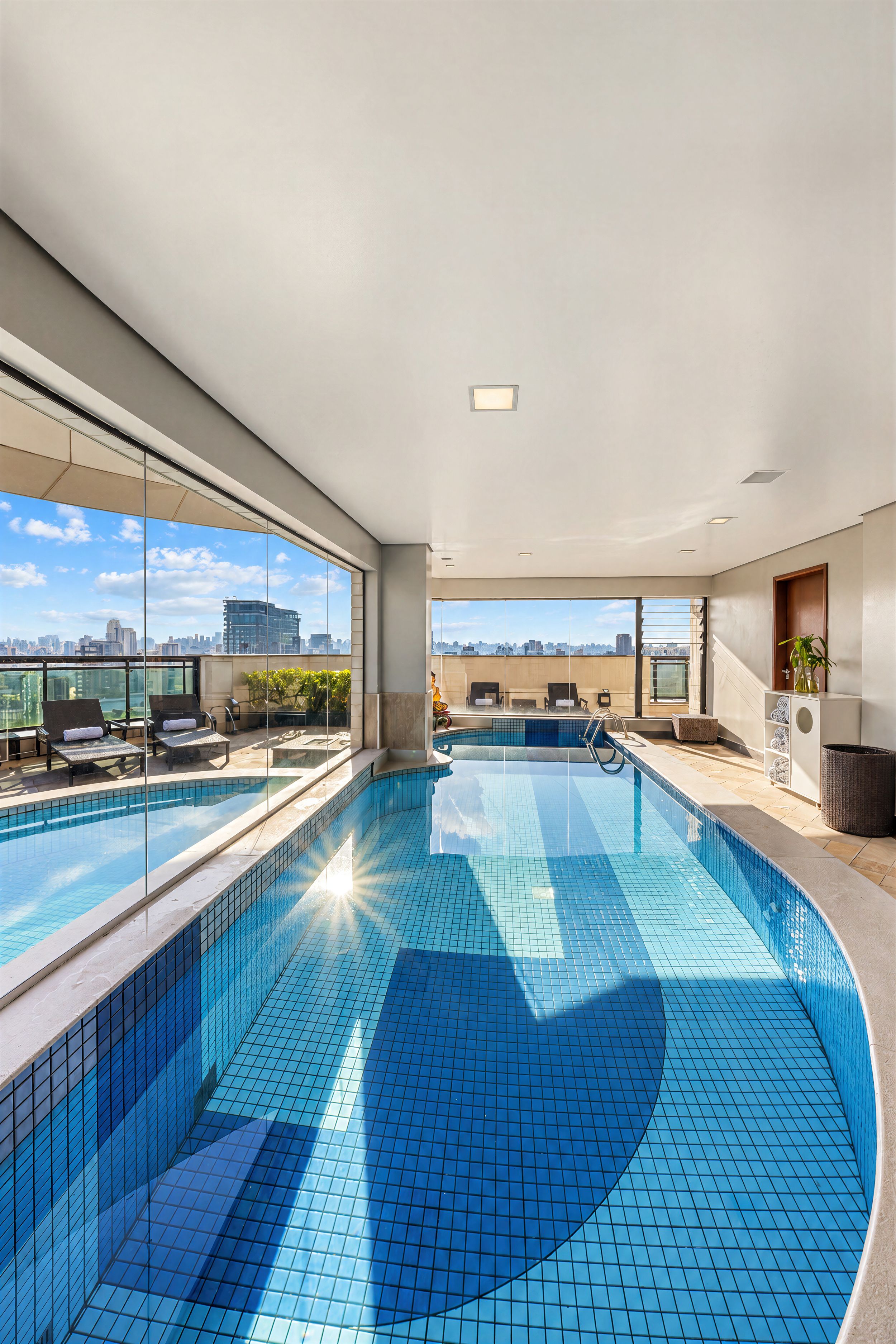 Una piscina interior moderna con paredes de vidrio transparente, vistas a la ciudad y una zona de piscina soleada.