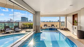 Una piscina interior moderna con paredes de vidrio transparente, vistas a la ciudad y una zona de piscina soleada.