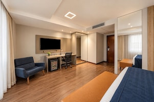 Quarto de hotel com cama, escrivaninha, cadeira, televisão e minibar.