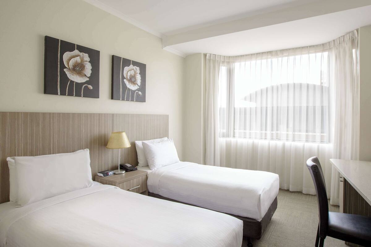 Ett hotellrum med två sängar, ett skrivbord, en stol, en lampa och ett fönster med gardiner.