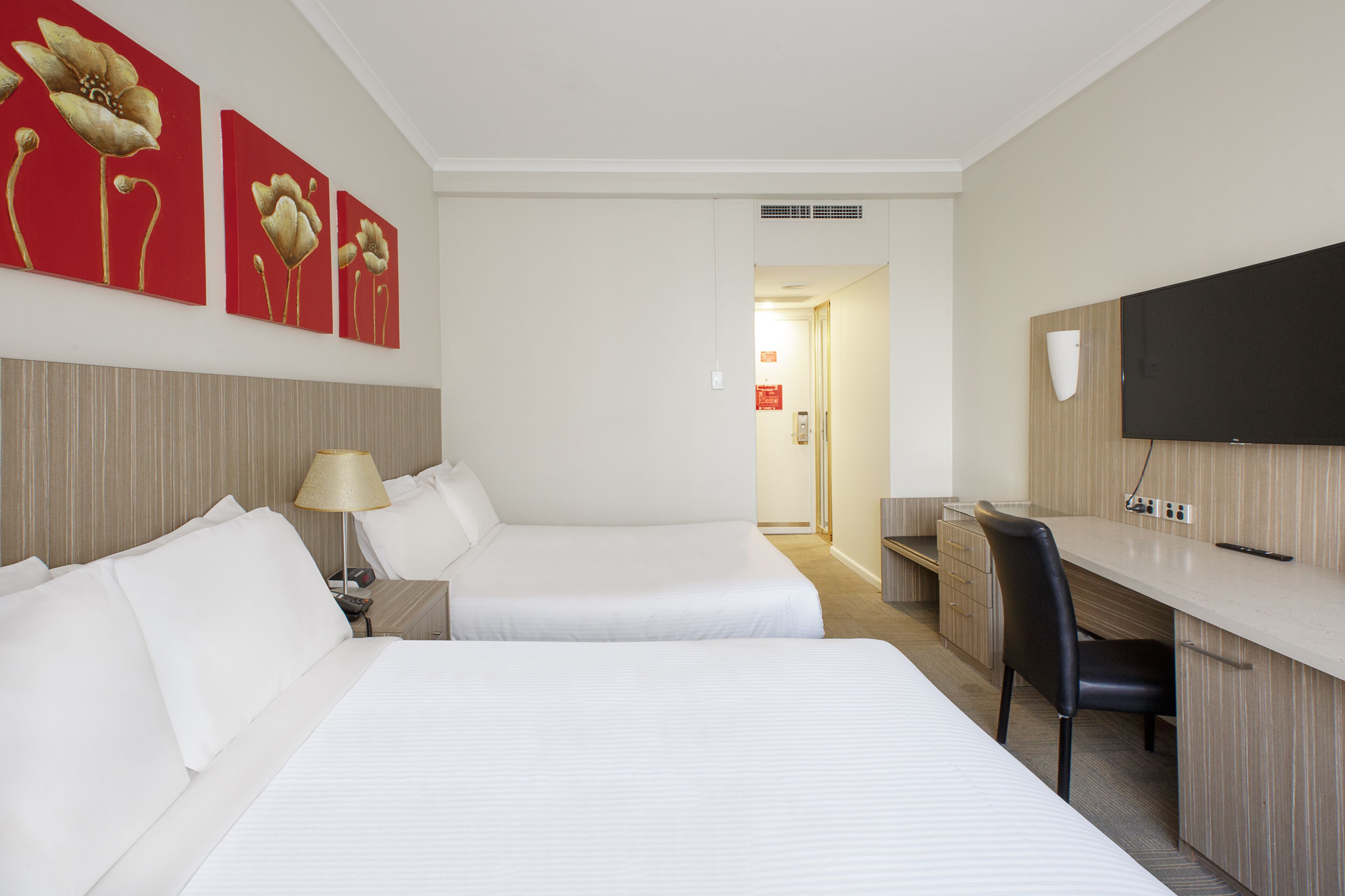 A hotel room with two beds, a desk, a chair, a TV, and artwork on the wall.
