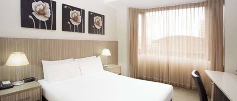 A hotel room with a large bed, two bedside tables with lamps, a desk, and a chair. There are three floral paintings on the wall.