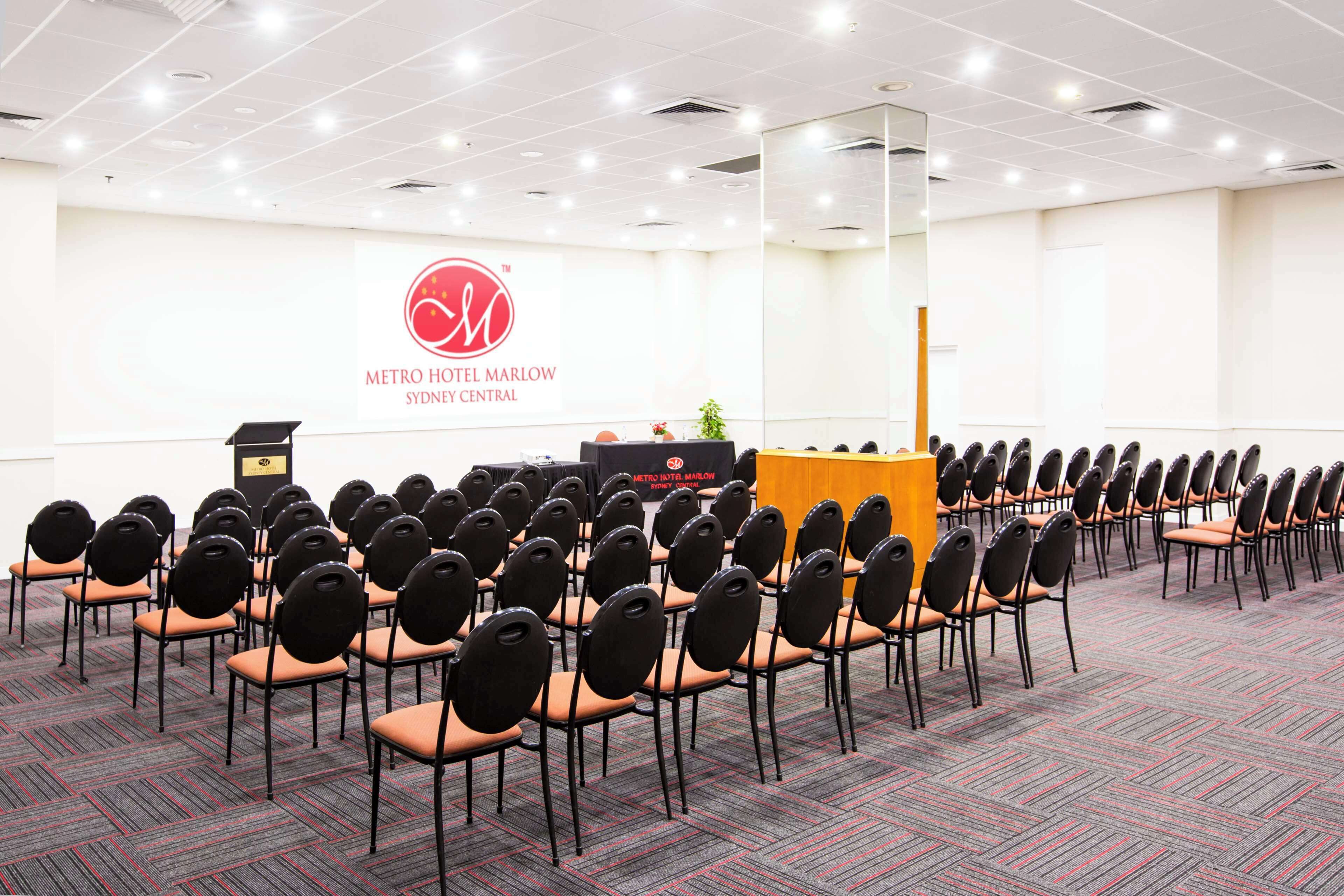 Une salle de conférence à l’hôtel Metro Mallow Sydney Central, avec des rangées de chaises, un pupitre et un grand logo sur le mur.