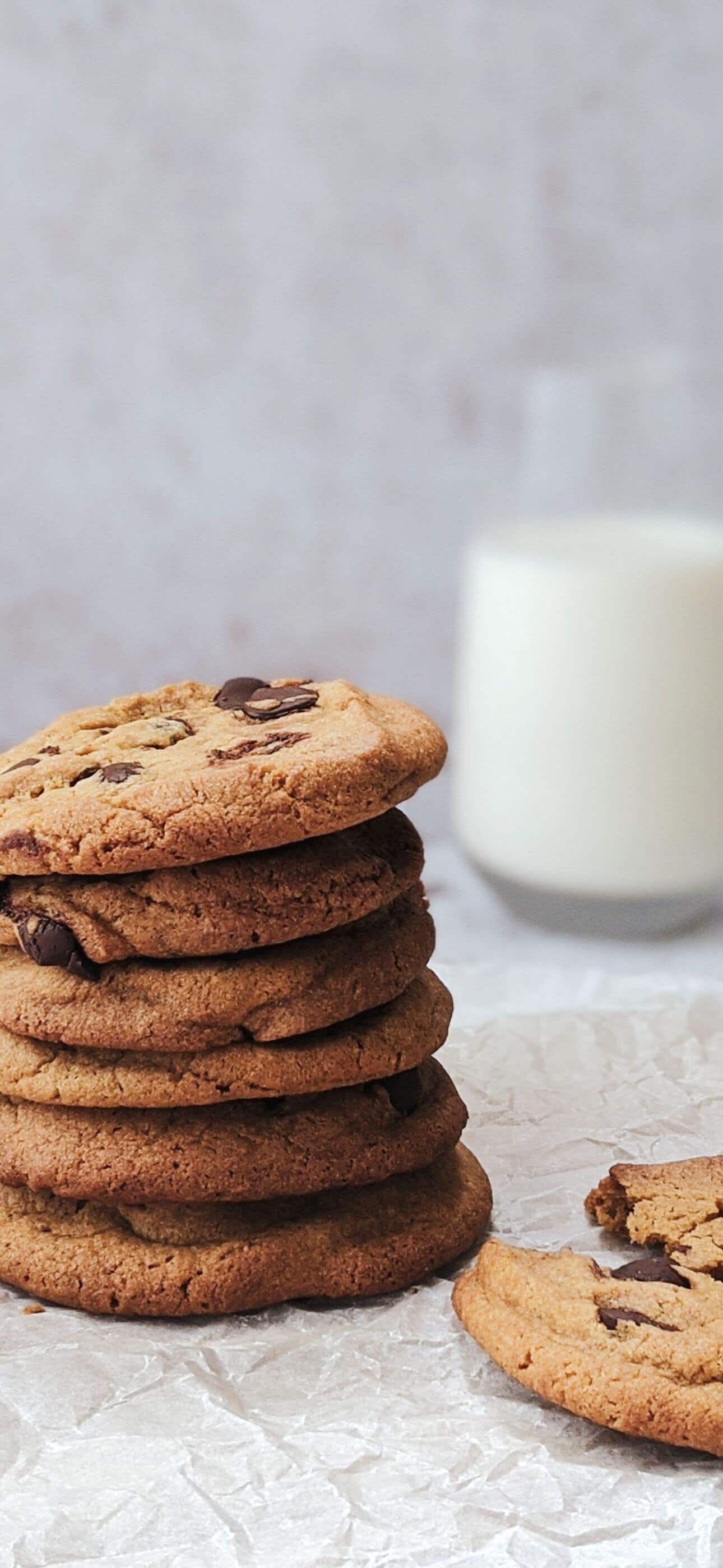 A stack of chocolate chip cookies on a crinkled paper surface with a glass of milk in the background.