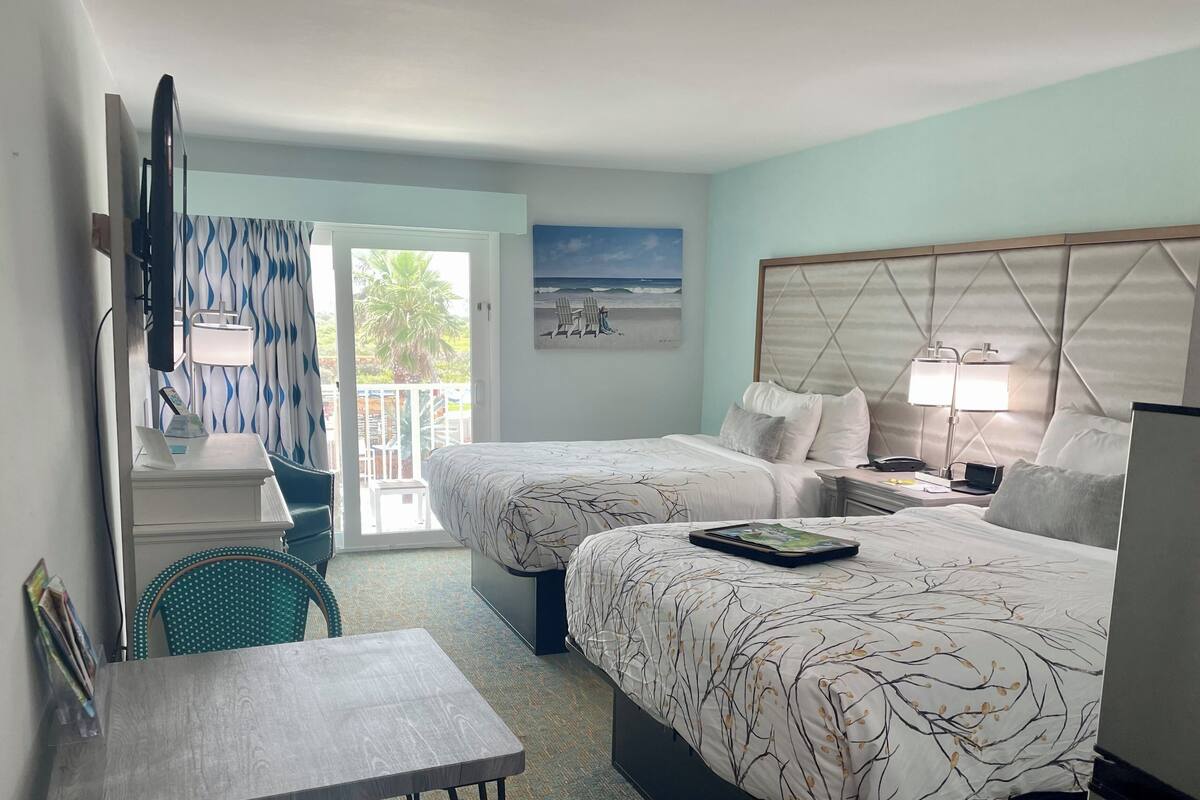Quarto de hotel com duas camas, uma escrivaninha, uma cadeira, uma televisão e um quadro de uma praia.